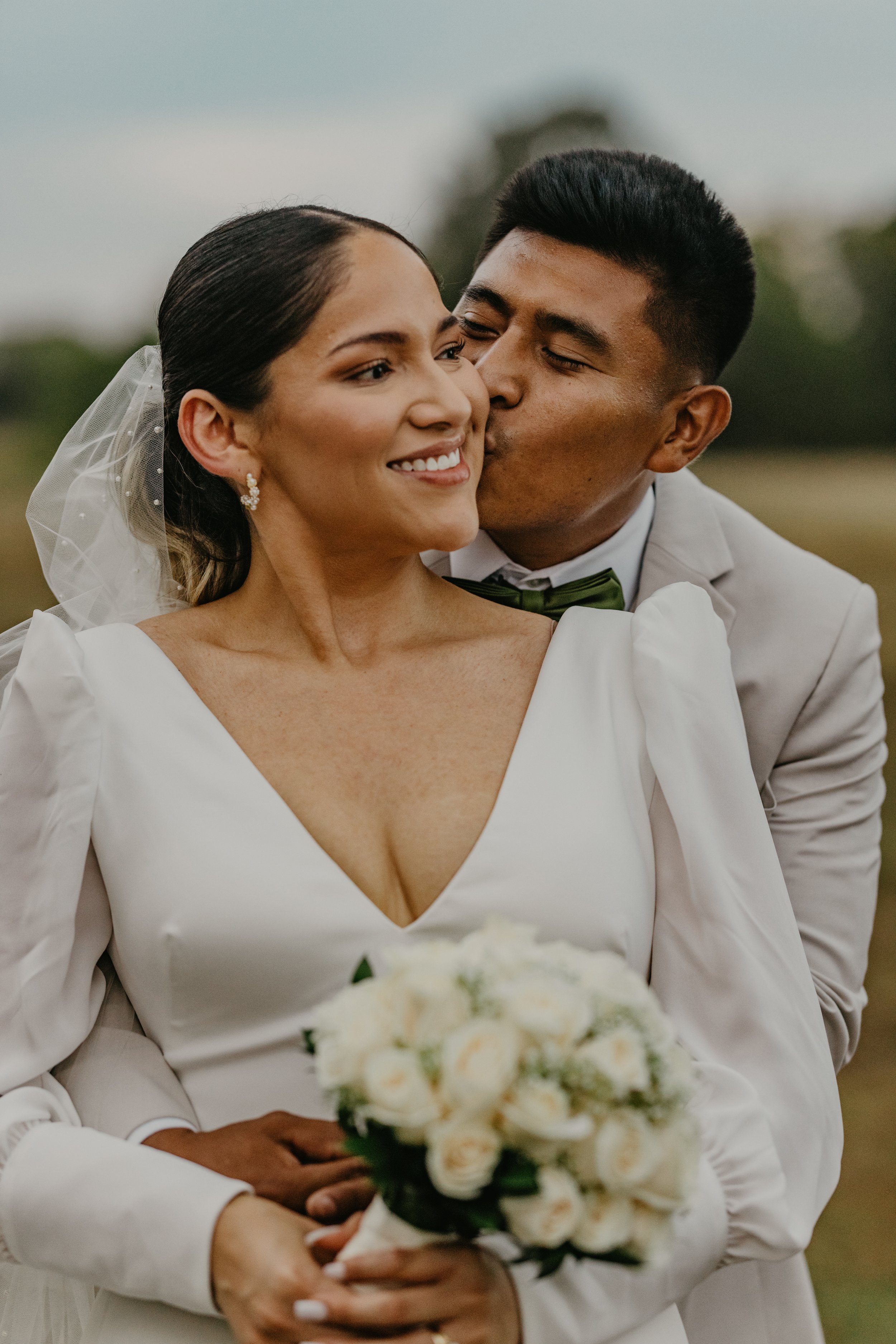 A bride and groom on their wedding day, with the groom kissing the bride's cheek while she holds a bouquet of white roses outdoors.