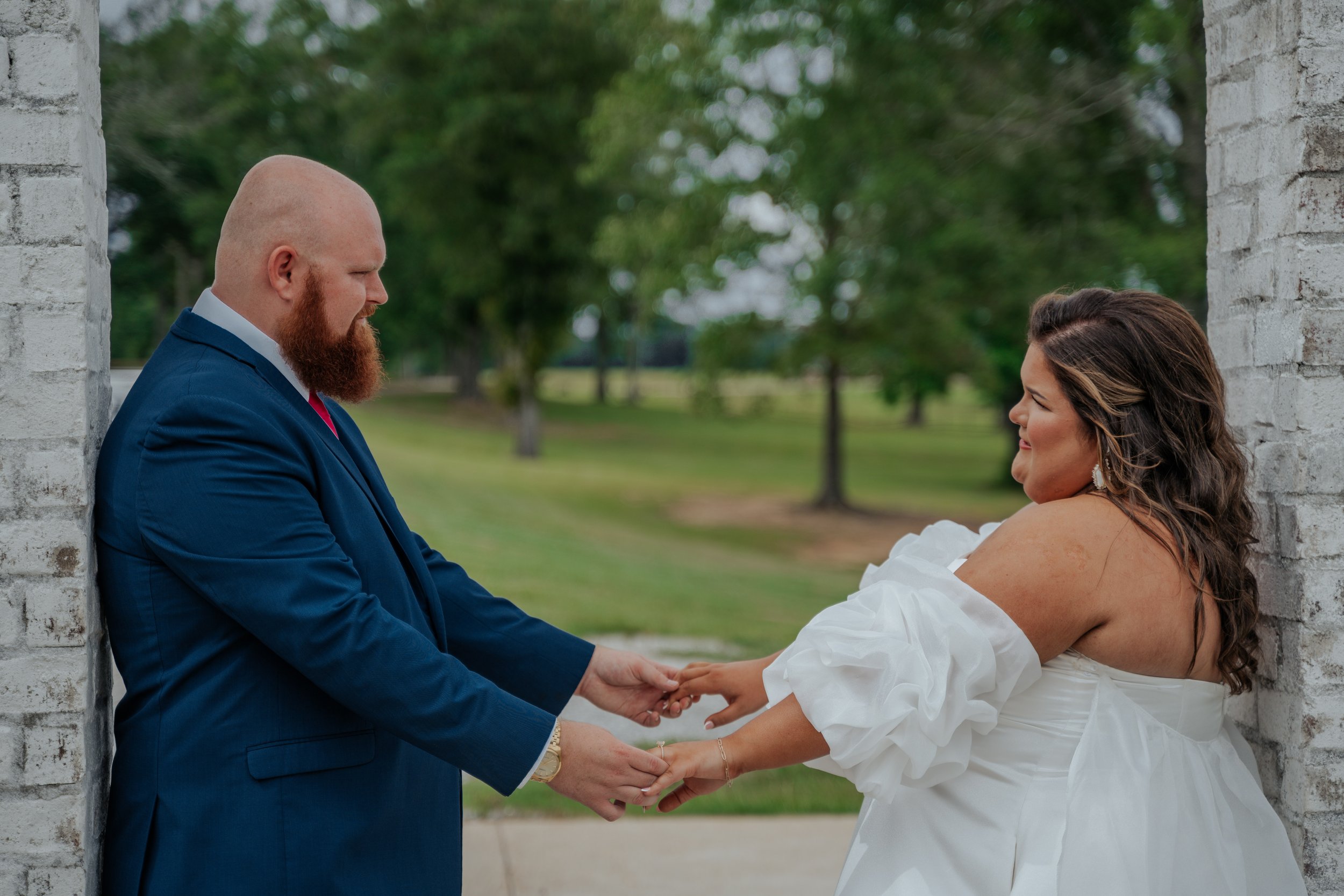 A couple exchanging rings outdoors, standing between two white brick columns, with a green park background.