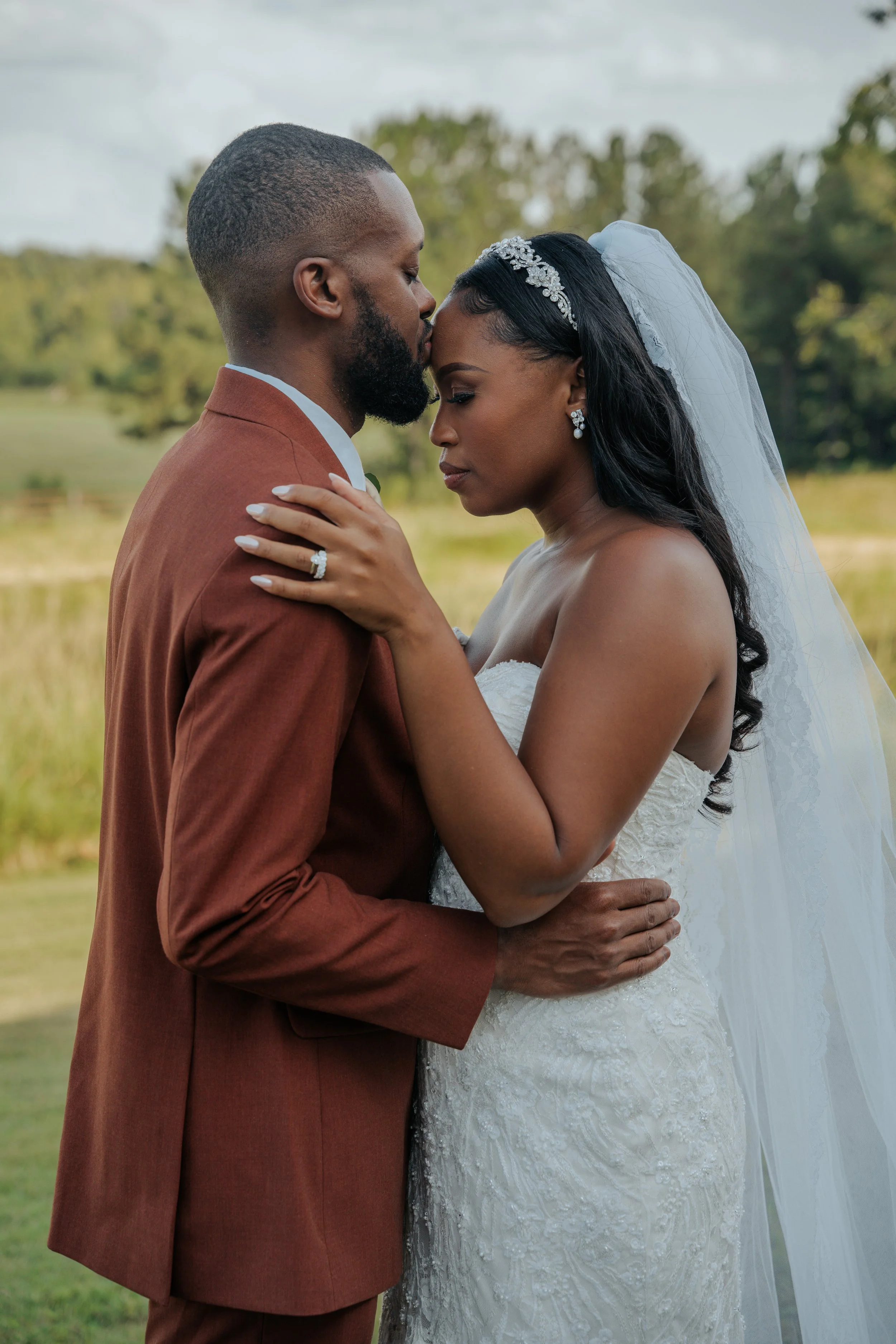 A bride and groom embrace outdoors, with the bride in a strapless white wedding gown and veil, and the groom in a brown suit, standing by a lake with greenery in the background.