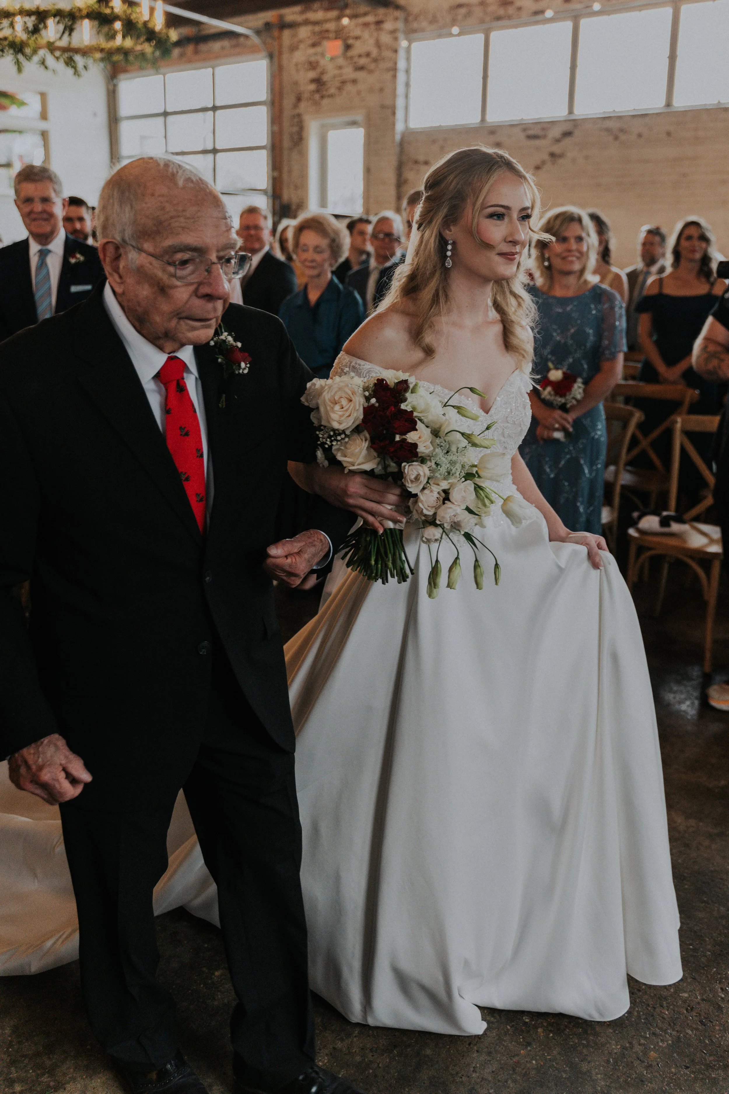 Bride walking down the aisle with her father during a wedding ceremony, holding a bouquet of flowers, in a rustic indoor venue with large windows and guests in the background.