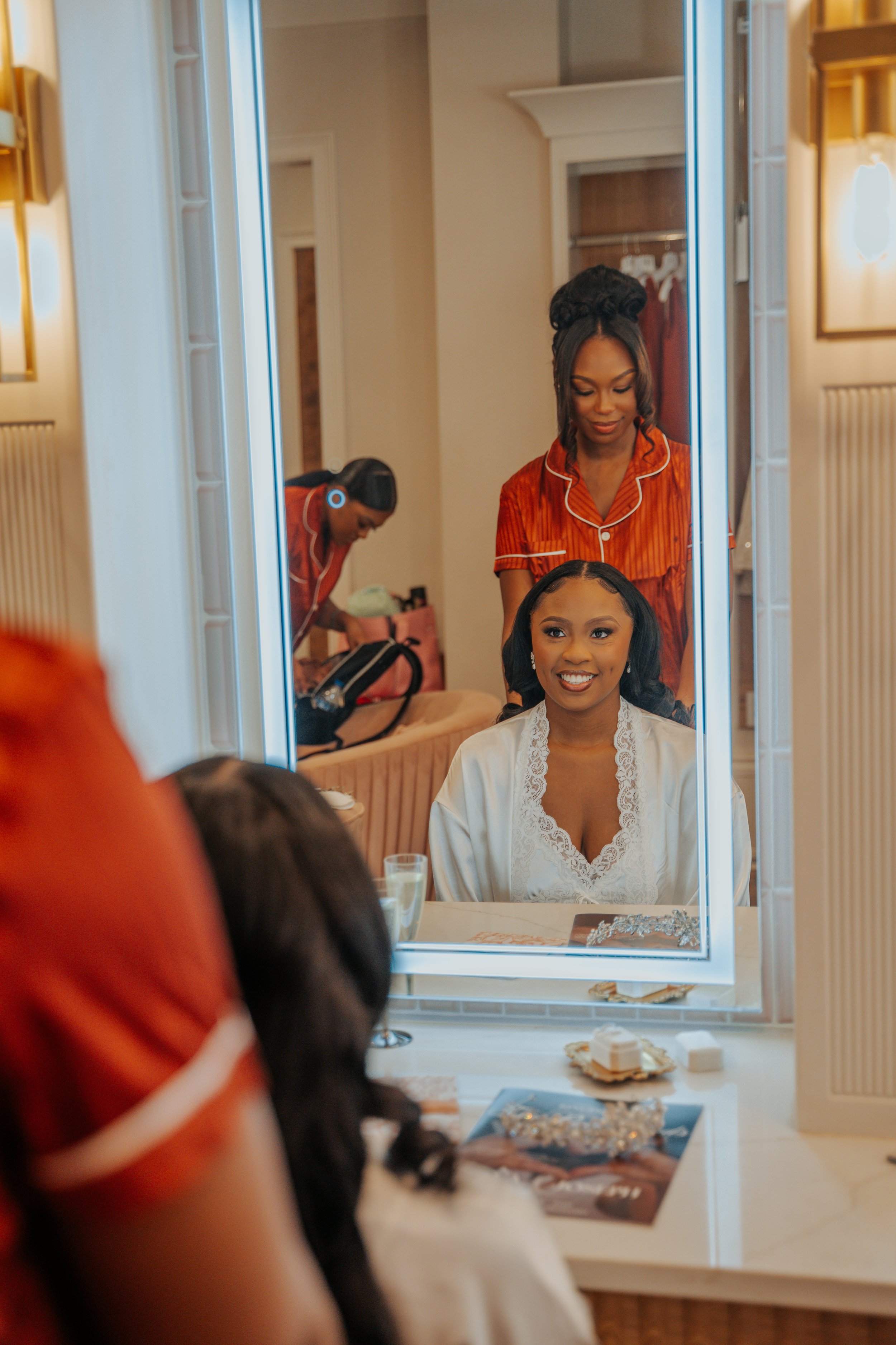 A woman getting her hair styled while looking into a mirror, with another woman behind her assisting, in a well-lit room with warm tones.