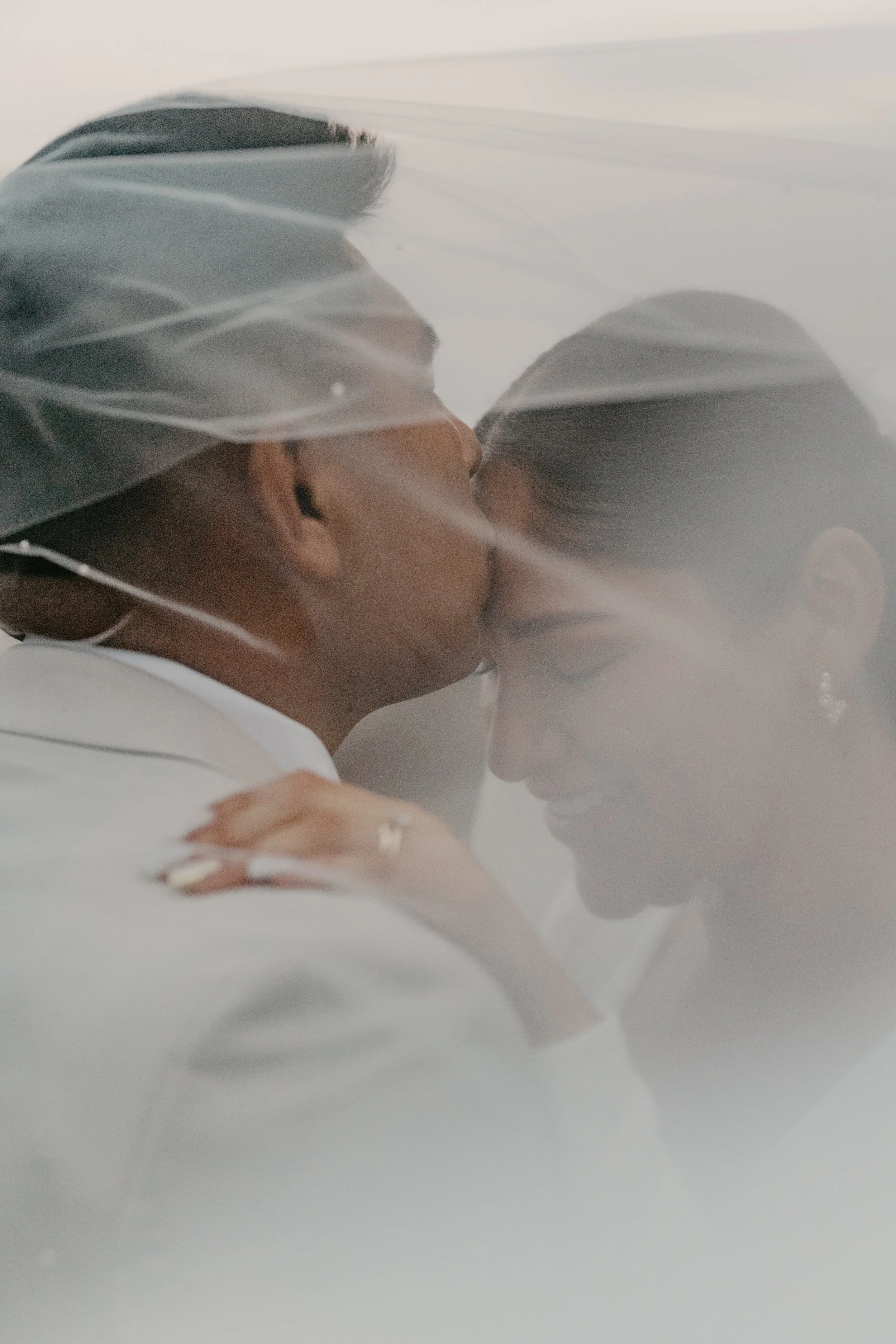 A bride and groom share a kiss behind a bridal veil, with soft lighting and a romantic atmosphere.