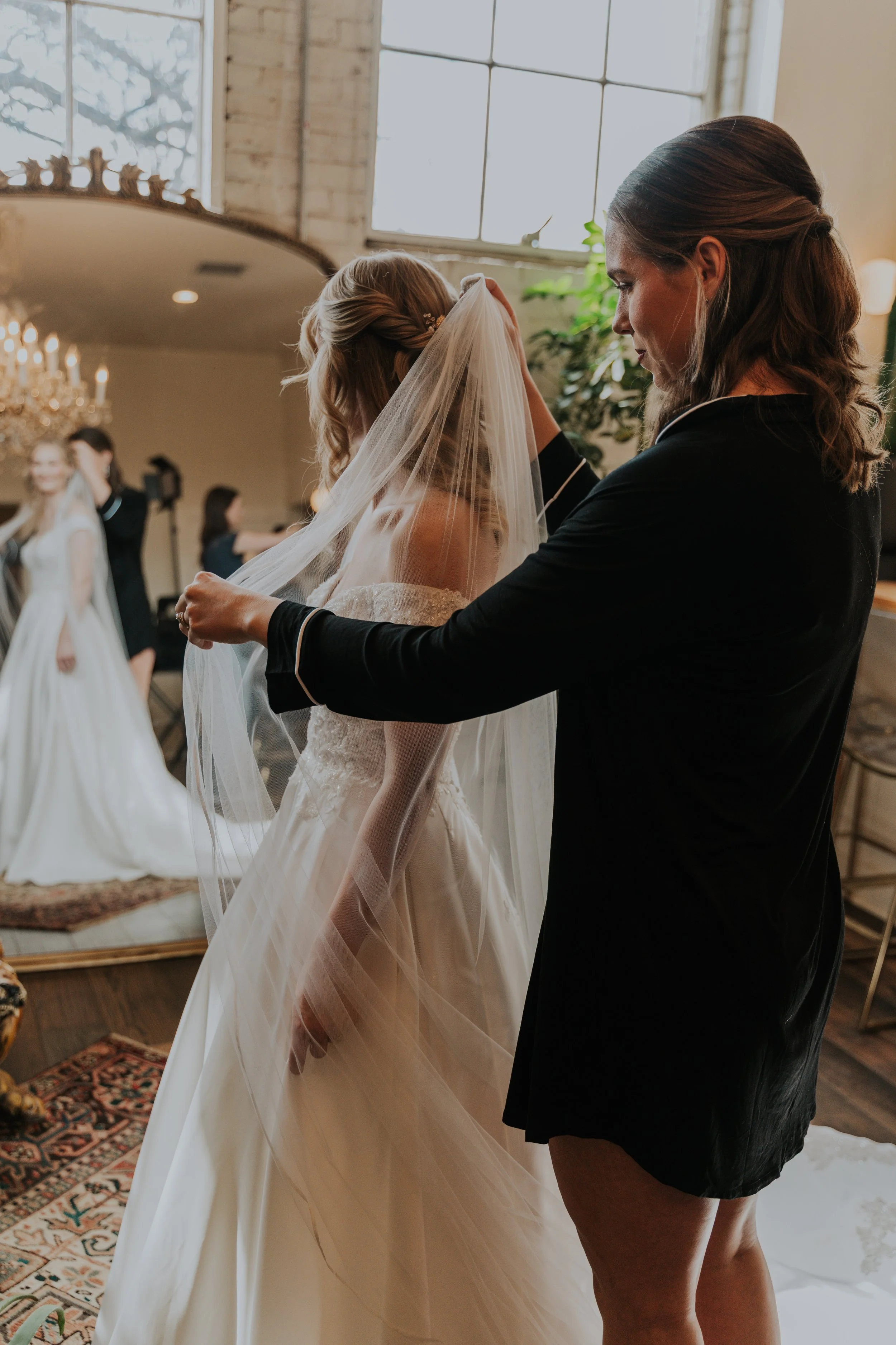 A bride in a wedding dress and veil is being helped by a woman in a black outfit in a warmly lit room with large windows, plants, and a chandelier, in preparation for her wedding.