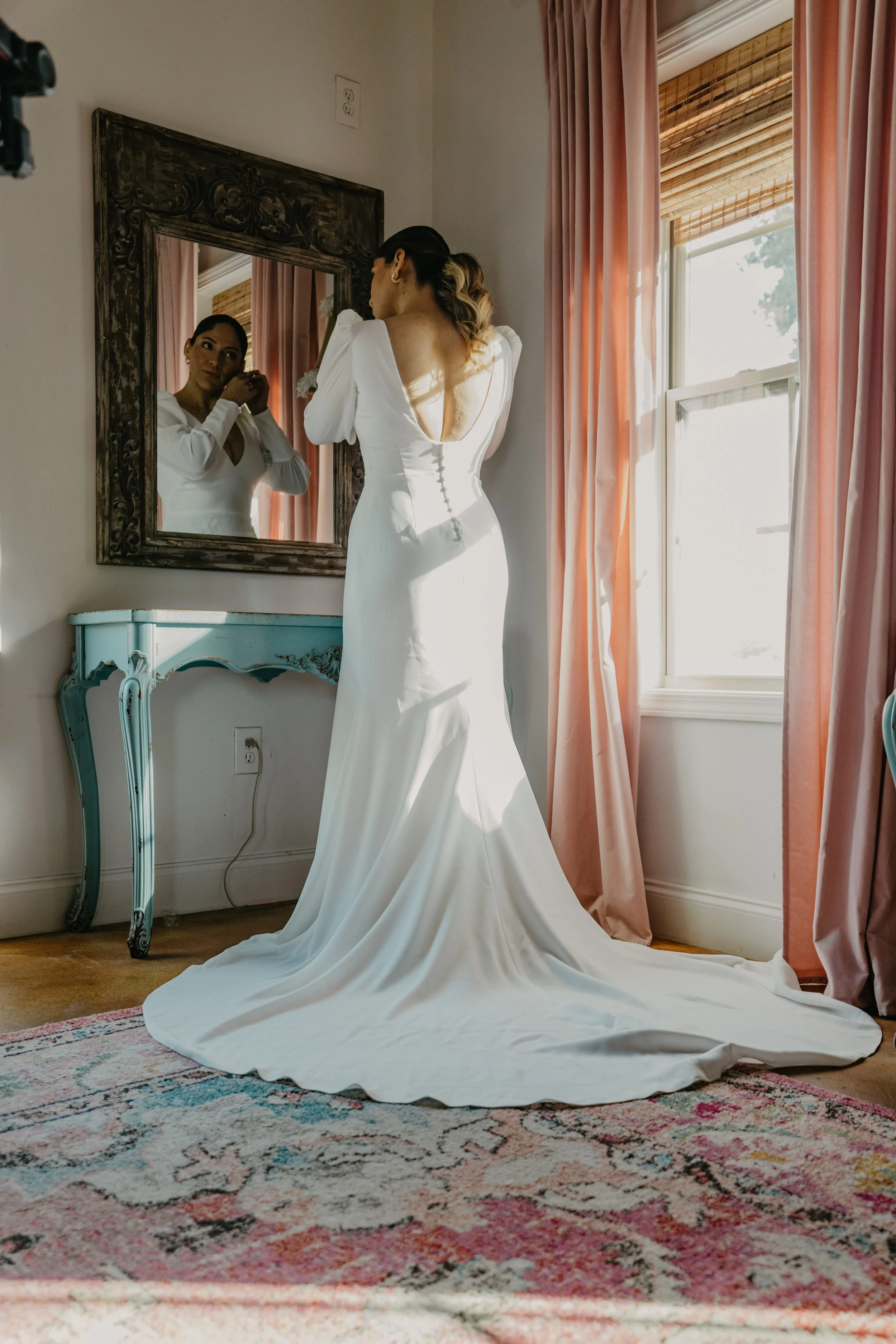 A woman in a white wedding dress stands in front of a mirror, adjusting earrings, with pink curtains and a window in the background.