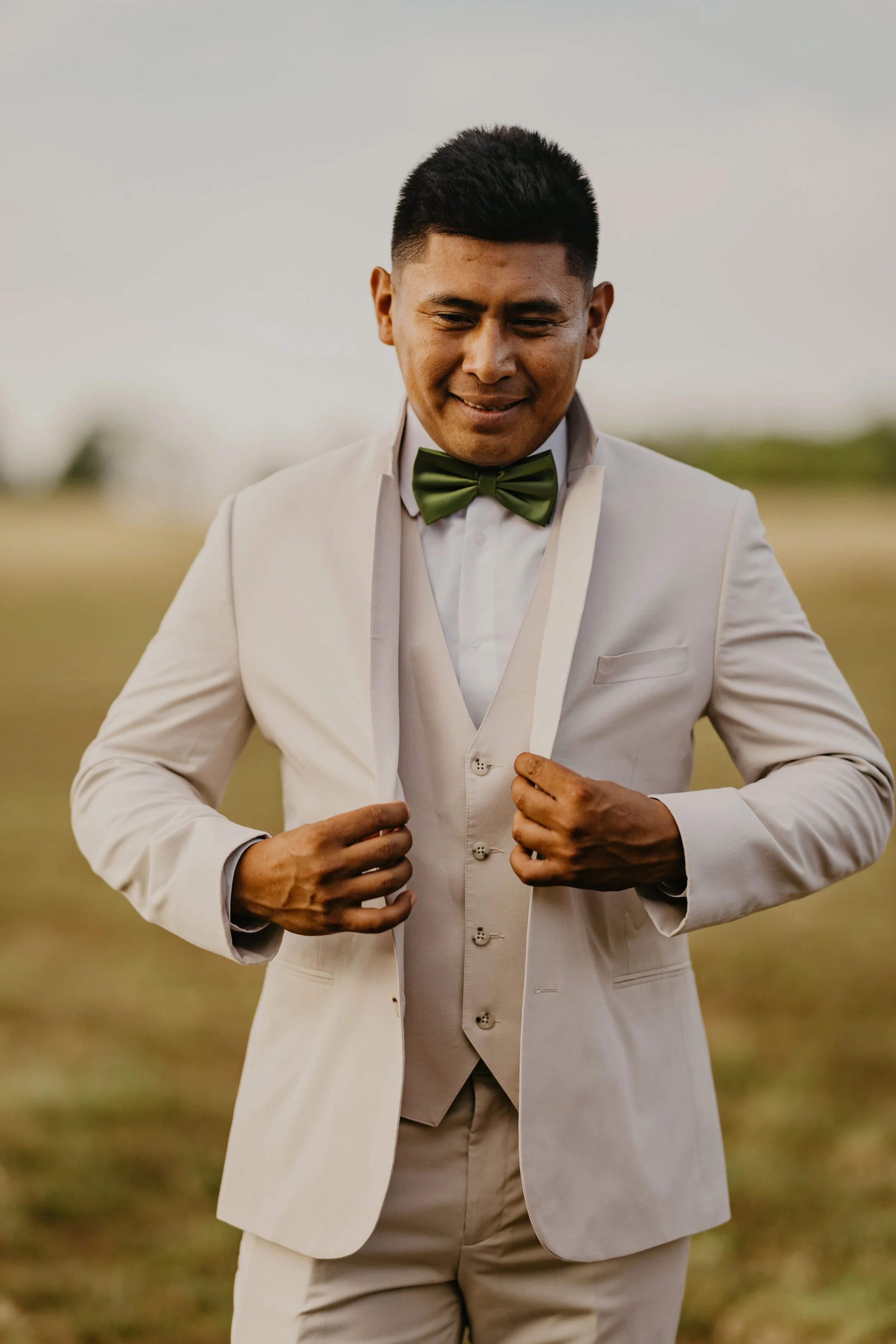 A man in a white suit with a green bow tie, adjusting his jacket outdoors.