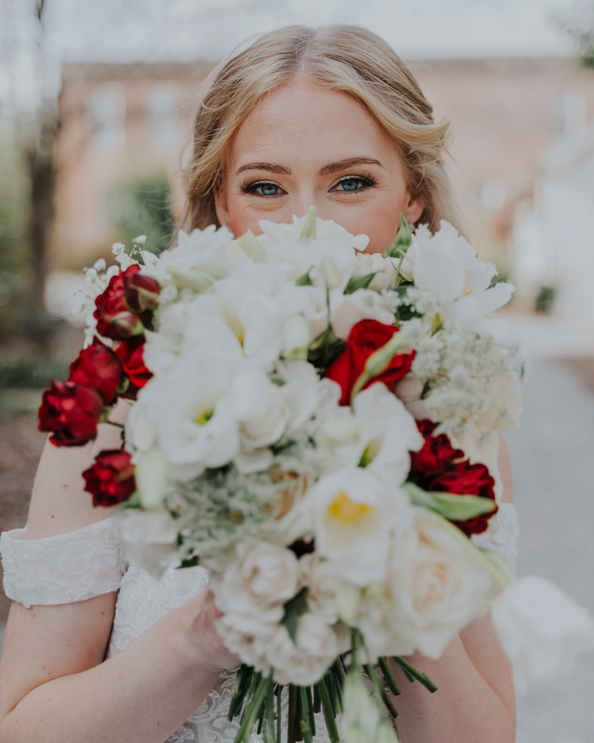 Bride holding a bouquet of white, red, and cream flowers in front of her face, outdoors.