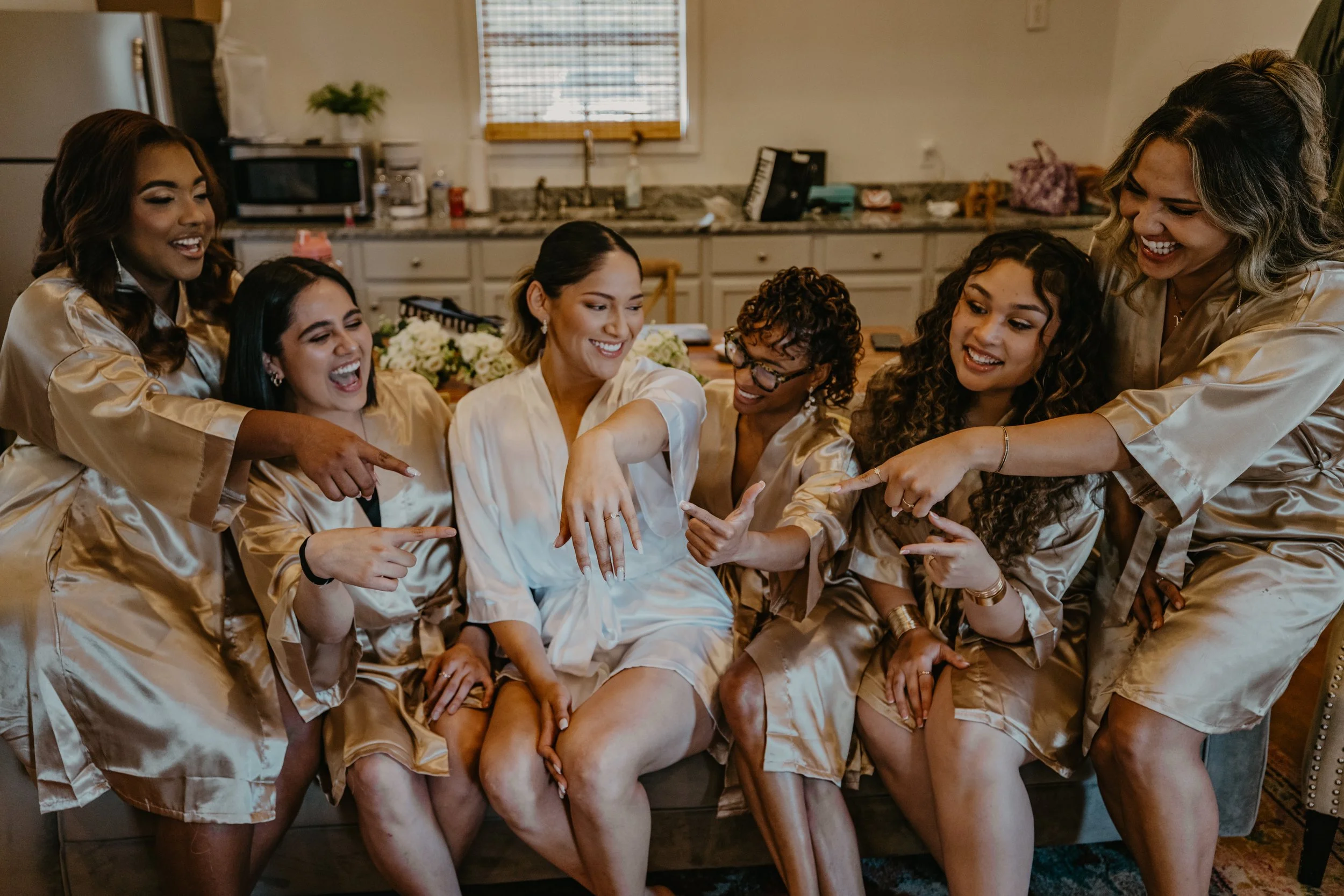 A group of seven women in matching satin robes, smiling and pointing at a woman's wedding ring, seated on a couch in a cozy kitchen.
