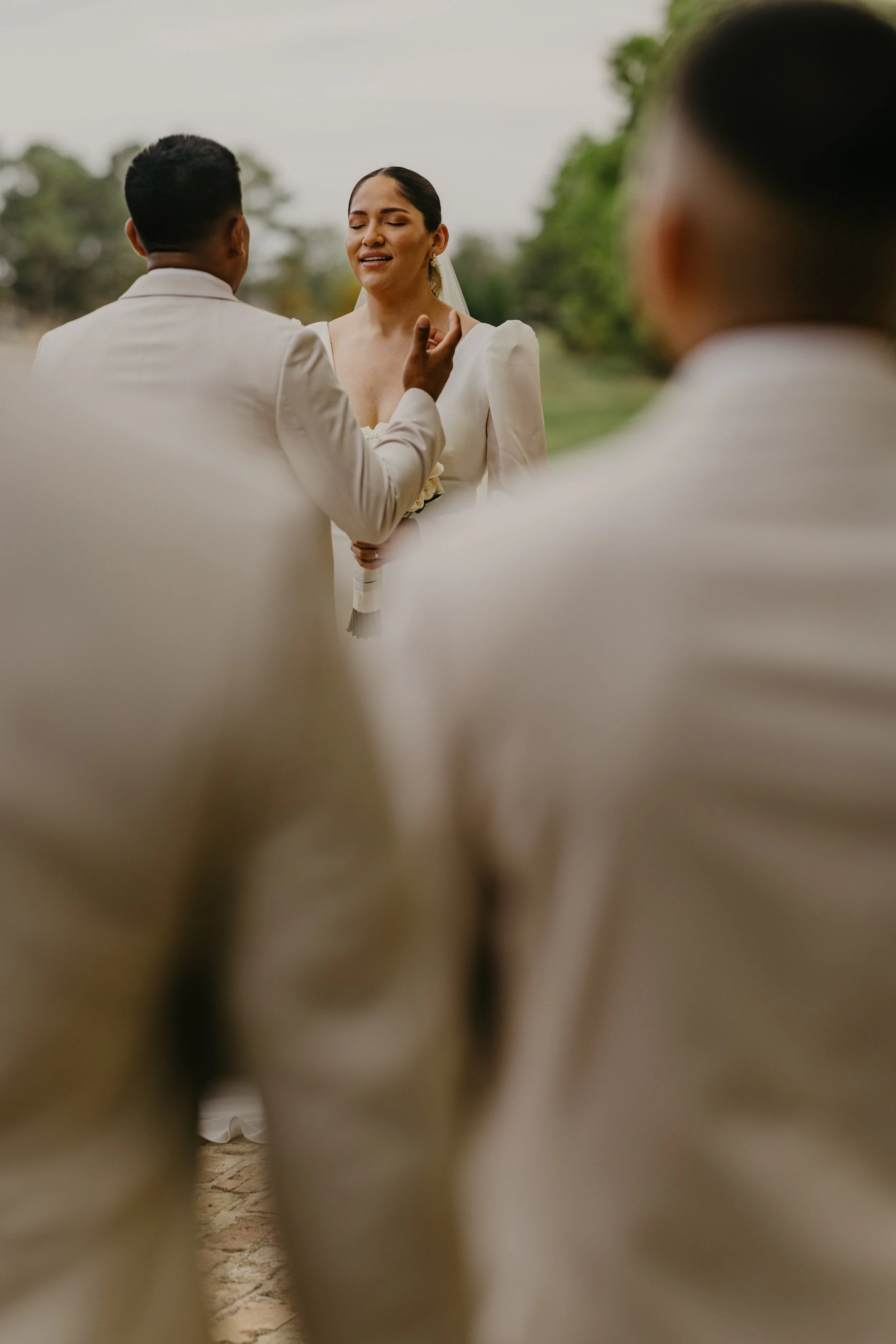 A woman in a wedding dress has her eyes closed and appears emotional as she says her vows, while a man in a white suit faces her and touches her chin during a wedding ceremony outdoors, with two other men in white suits in the foreground.