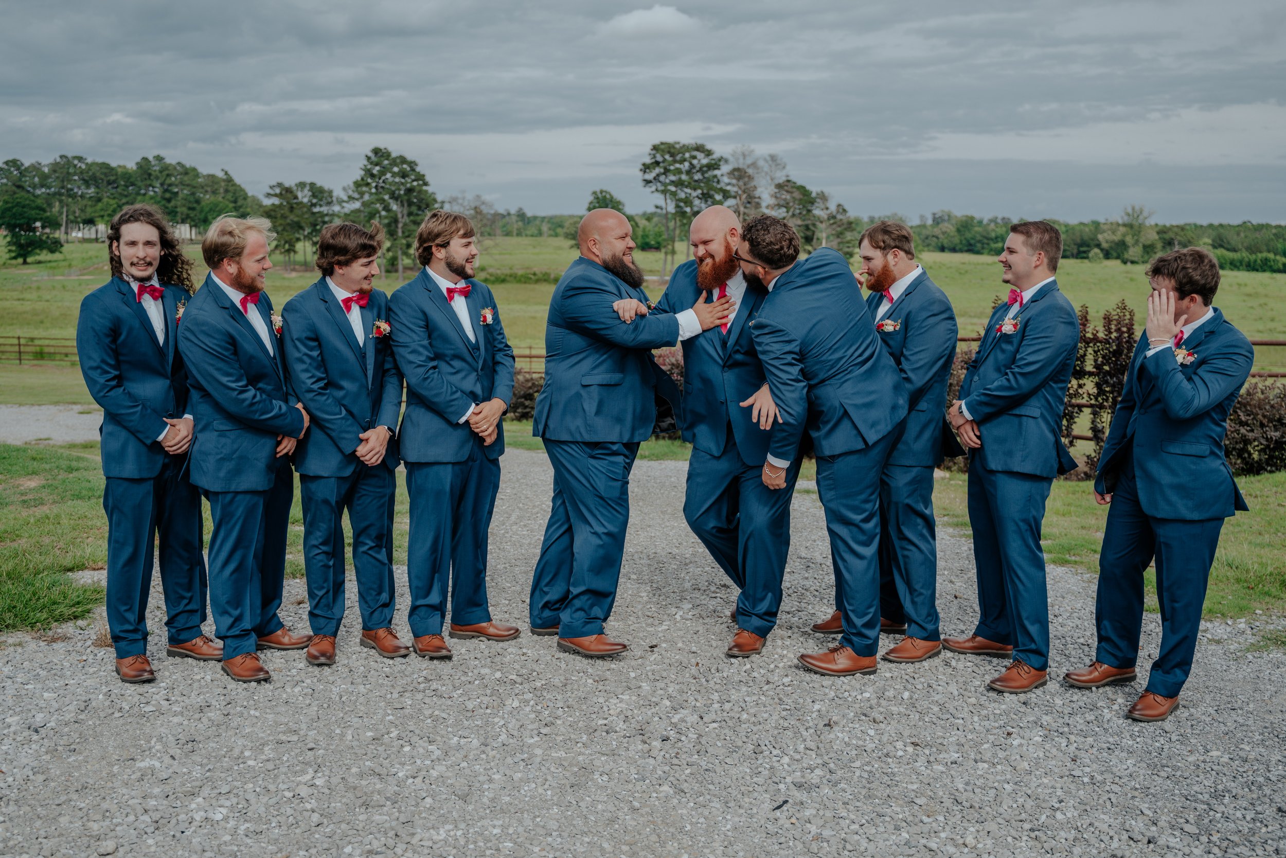 A group of nine men in matching blue suits and red bow ties, with some wearing boutonnières, laughing and playfully interacting outdoors on a gravel path, with a green landscape and cloudy sky in the background.