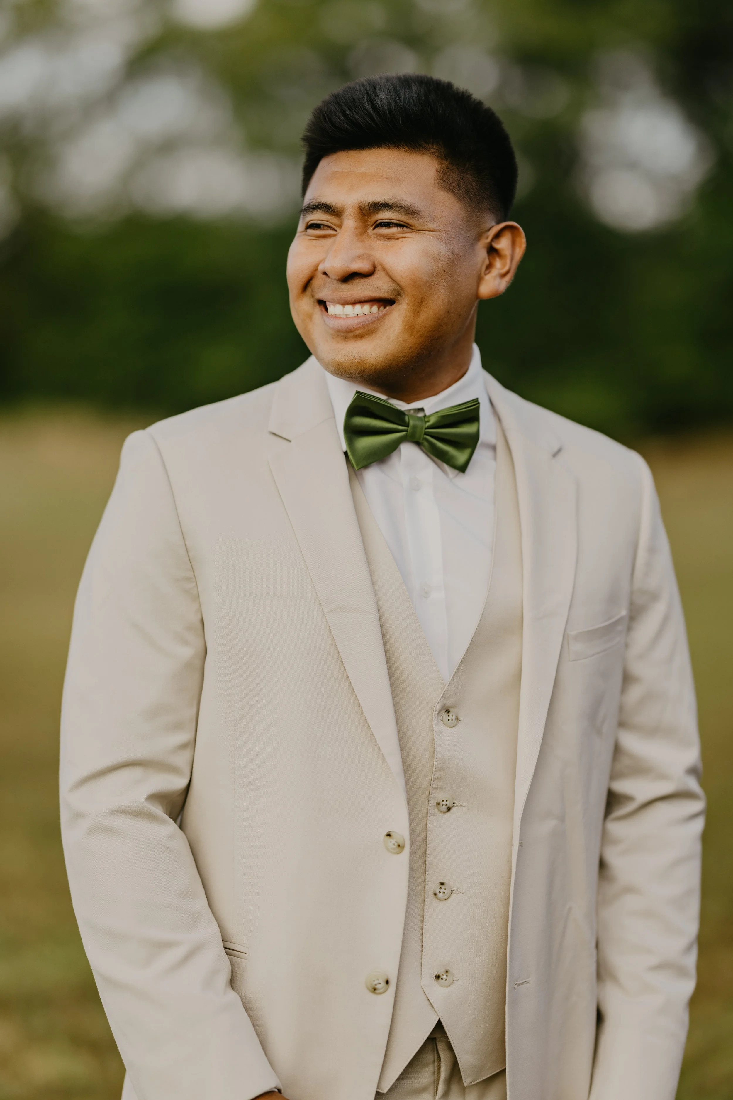 A man in a light beige suit with a matching vest, white shirt, and green bow tie, smiling outdoors in a natural setting with blurred greenery background.