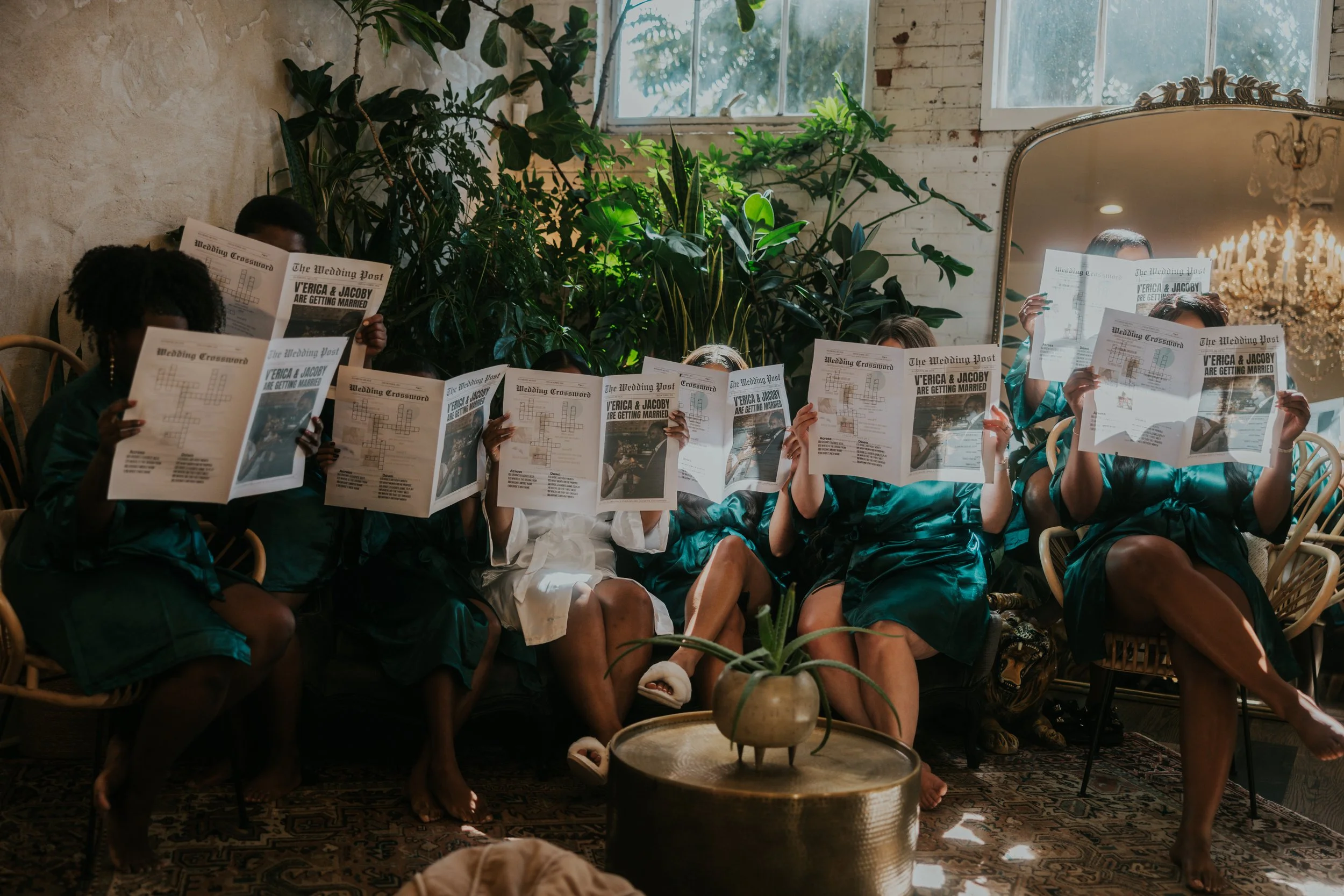 Group of women sitting on a couch, reading newspapers with wedding announcements inside a sunlit room with plants, a chandelier, and a large mirror.