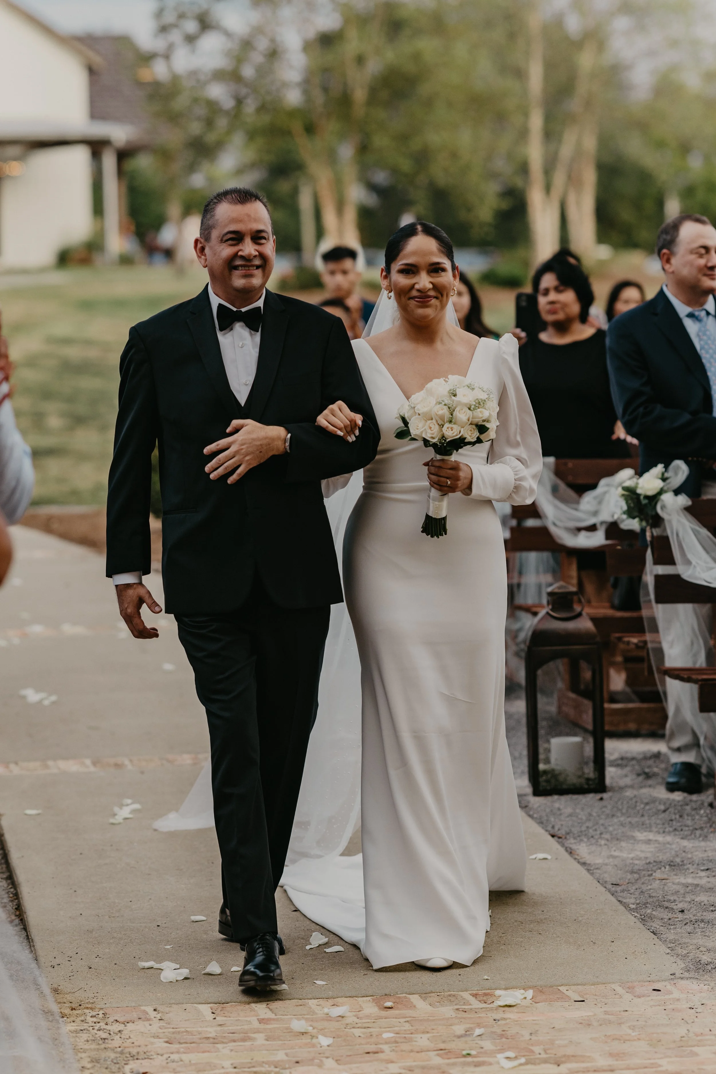 A bride in a white wedding dress walking arm-in-arm with a man in a tuxedo outdoors during a wedding ceremony, with guests seated nearby and trees in the background.