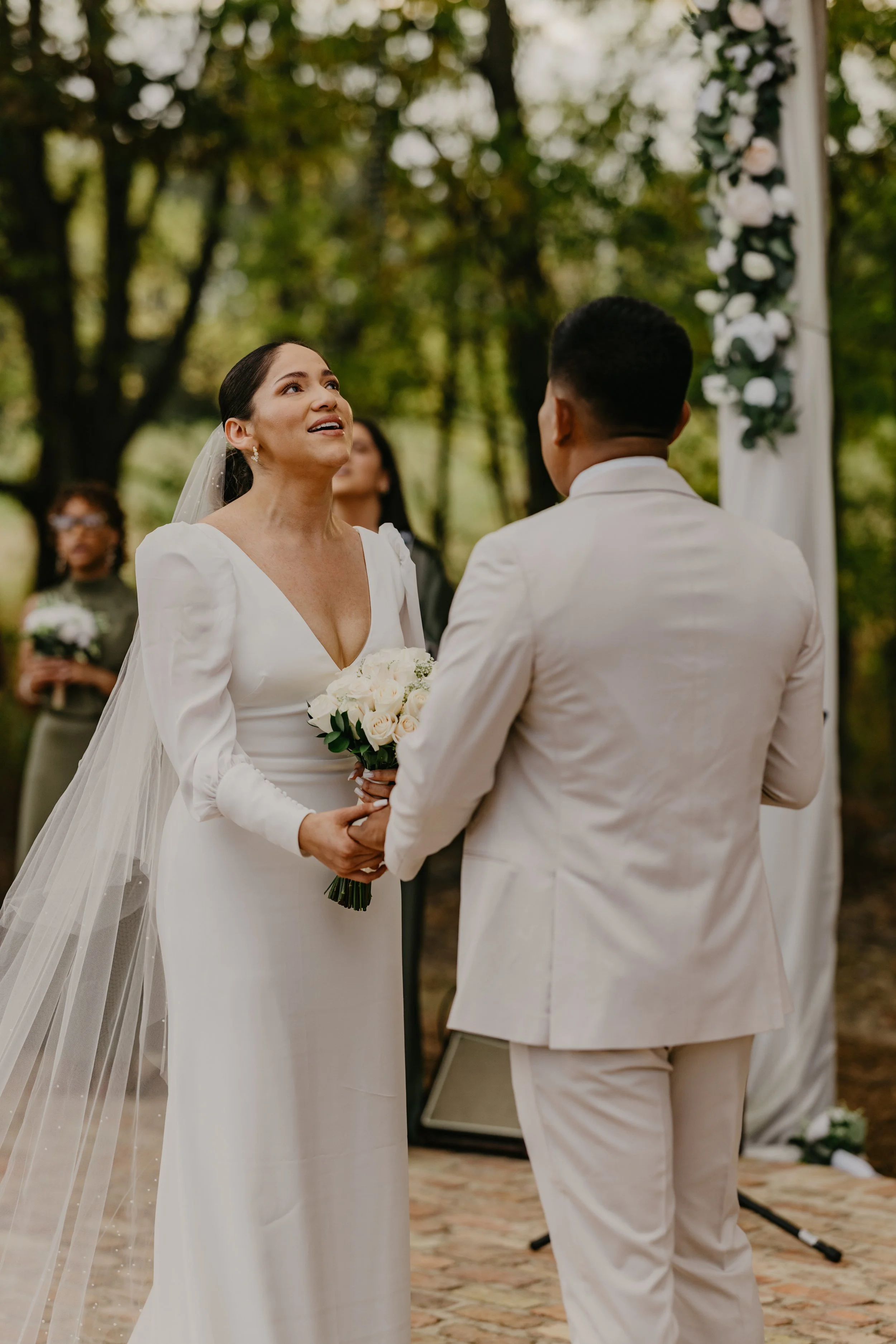 A bride and groom exchanging vows during an outdoor wedding ceremony, with the bride holding a bouquet of white roses and a floral arch decorated with white flowers in the background.