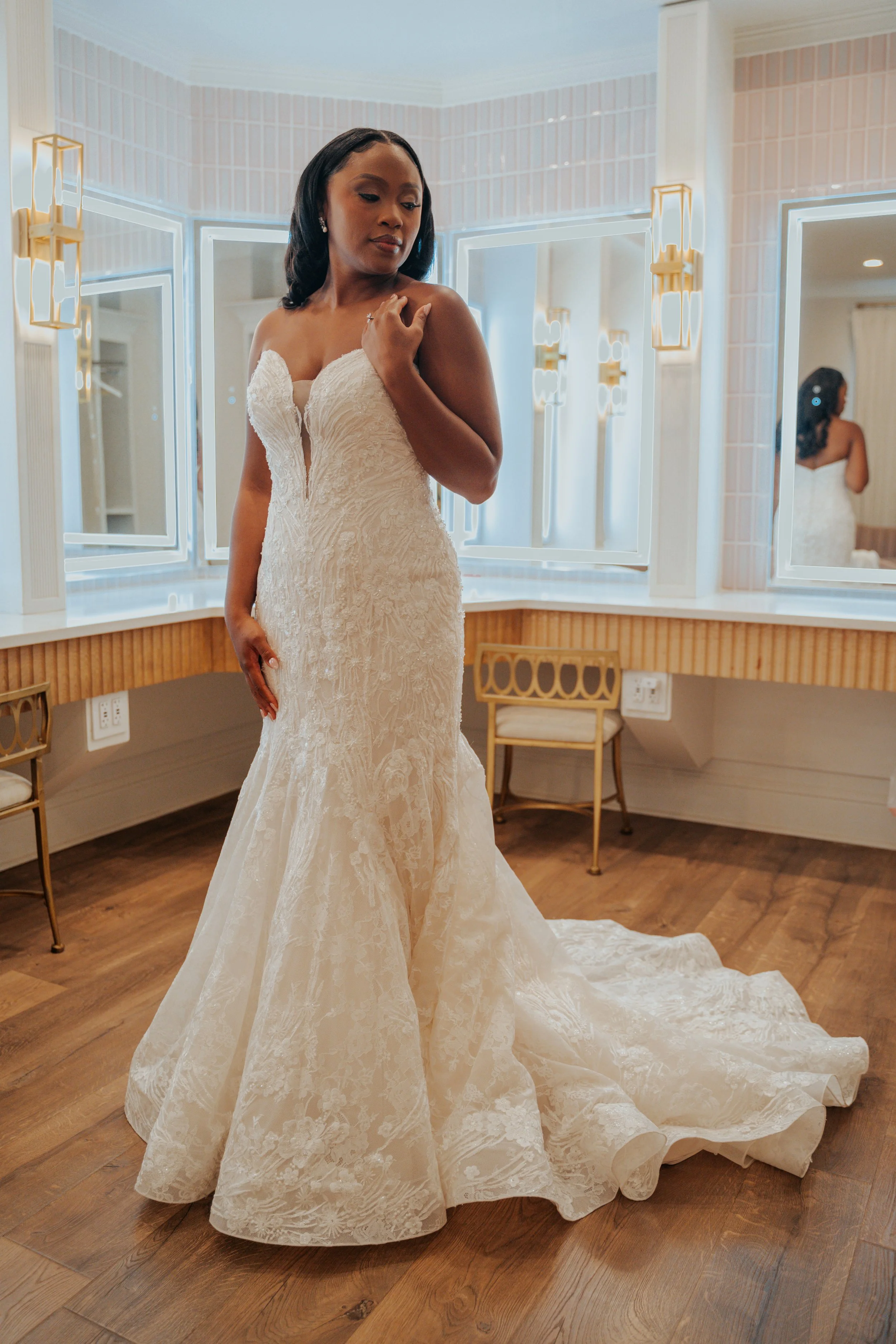 A woman in an elegant white lace wedding dress is standing in a bright dressing room with multiple mirrors, looking down. The room has wood flooring, gold and white chairs, and modern wall sconces.