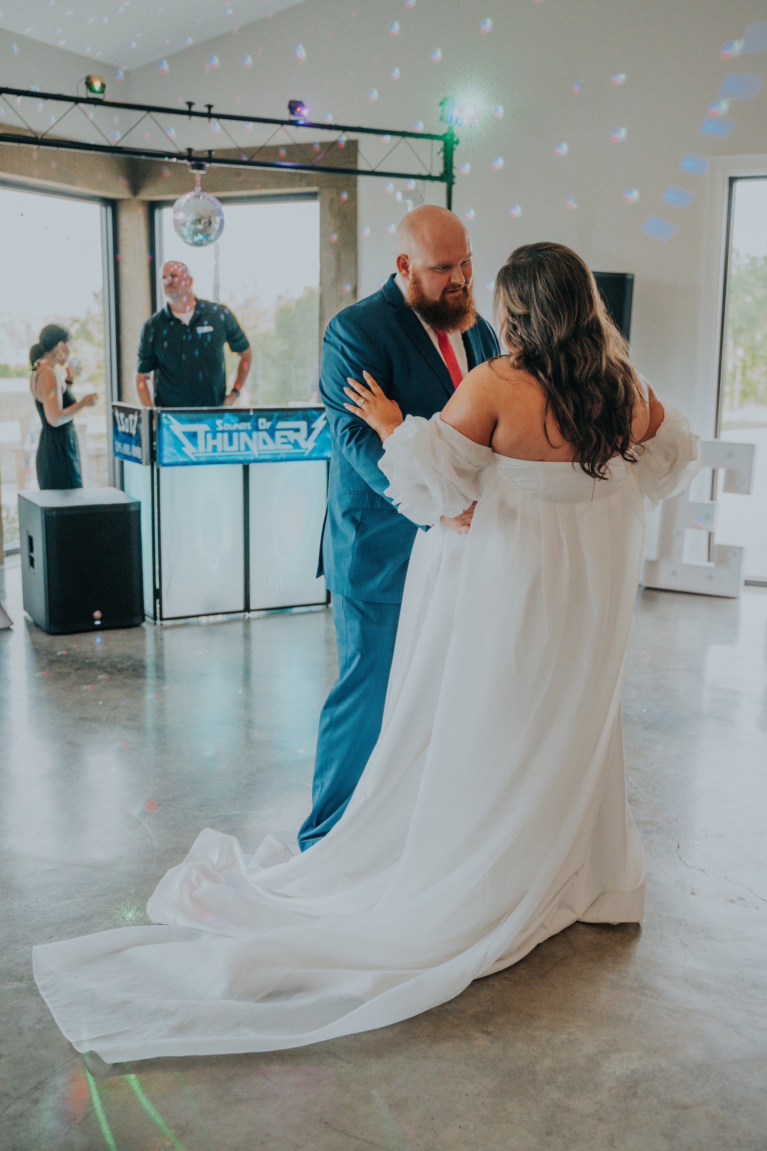 A bride and groom dancing at their wedding reception, with a DJ and guests in the background.