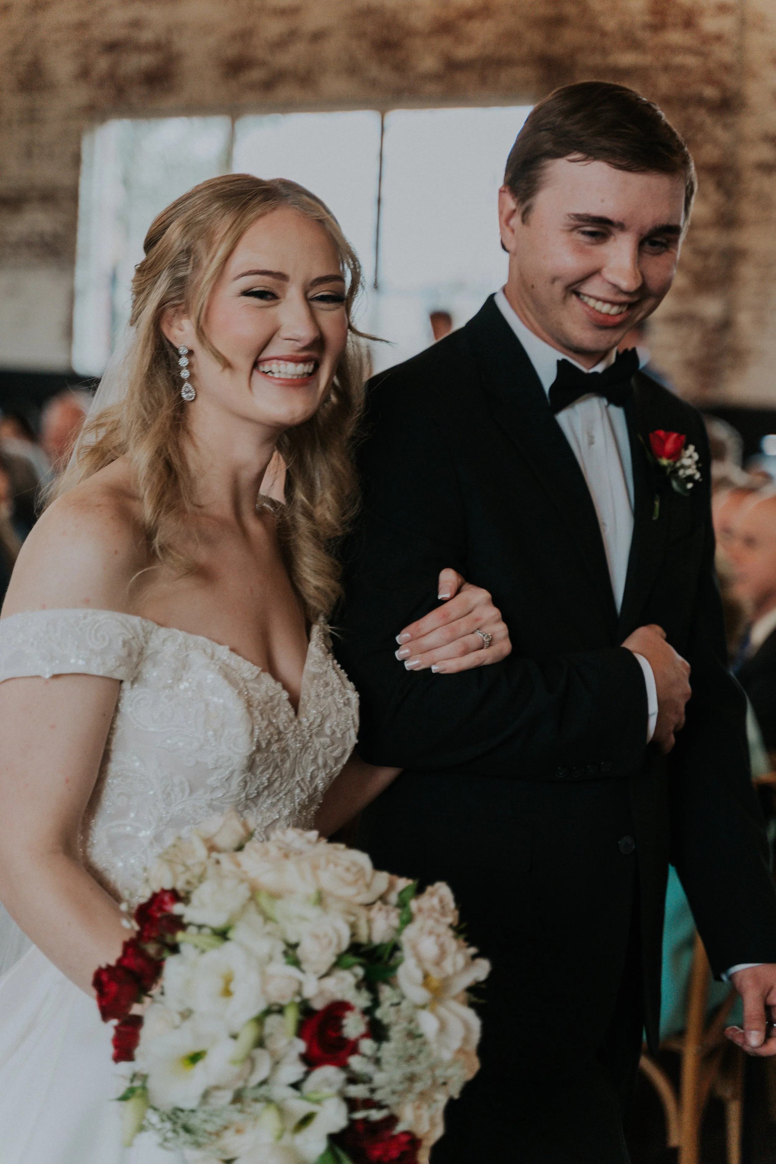 A bride and groom smiling as they walk down the aisle during their wedding ceremony. The bride is holding a bouquet of flowers and wearing a wedding dress, while the groom is dressed in a black tuxedo with a red boutonniere.