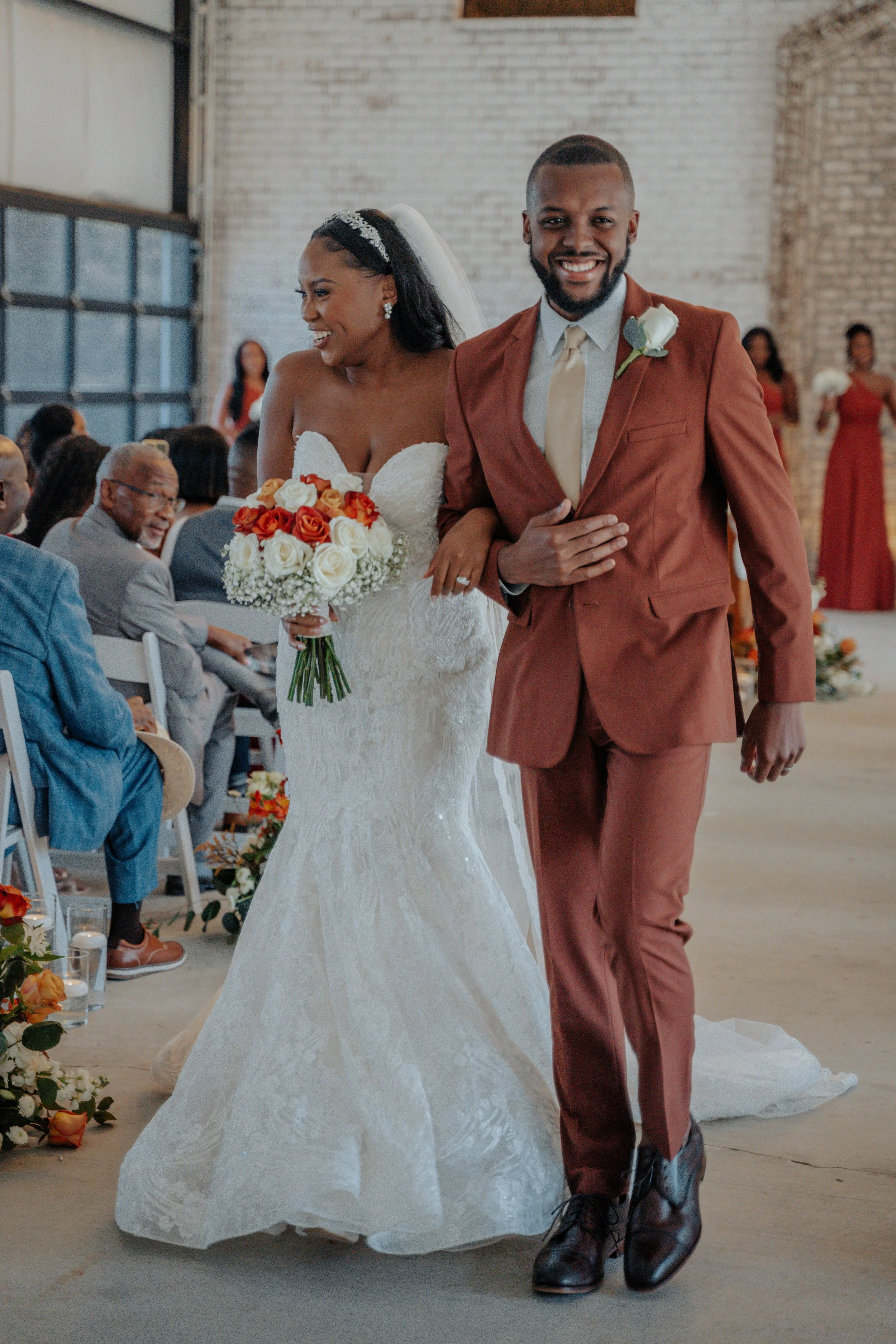A newlywed couple walking down the aisle at their wedding ceremony, with the bride holding a bouquet of white and red roses, and the groom smiling with his hand on his chest.
