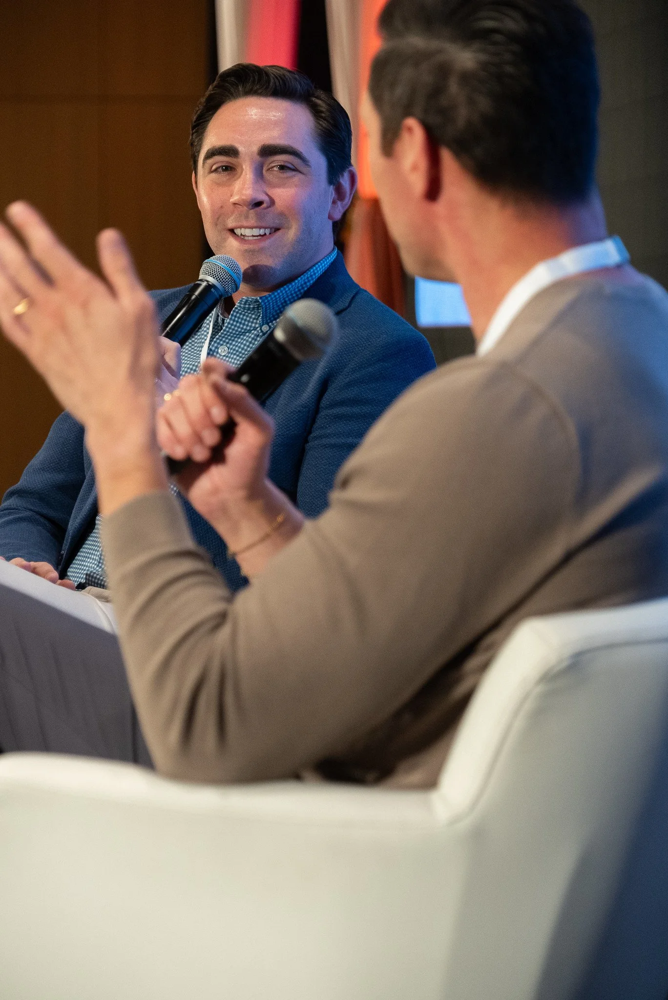 Two men are sitting and speaking into microphones during a discussion or interview, with one man looking at the other