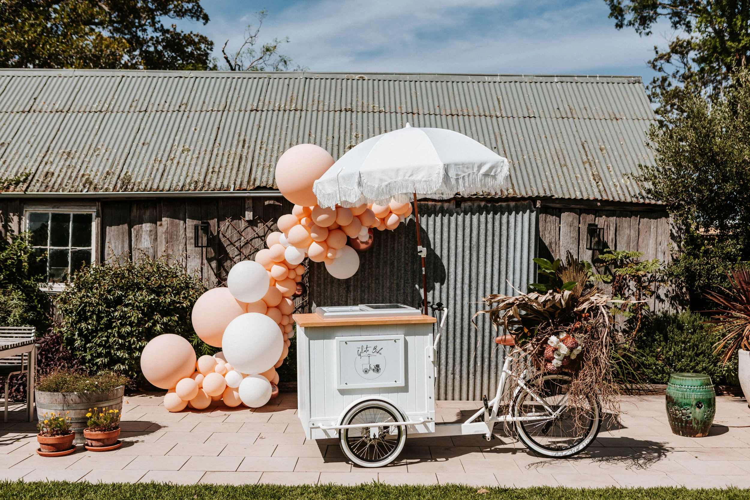 White gelato bike with white umbrella and balloon garland for wedding dessert catering