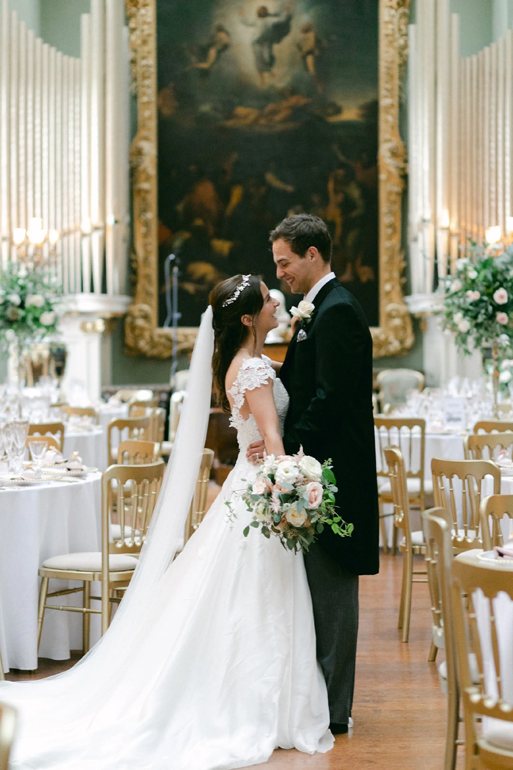 A bride and groom smiling and looking at each other at their wedding reception in a decorated hall with tables, chairs, and floral arrangements, with a large painting hanging on the wall behind them.