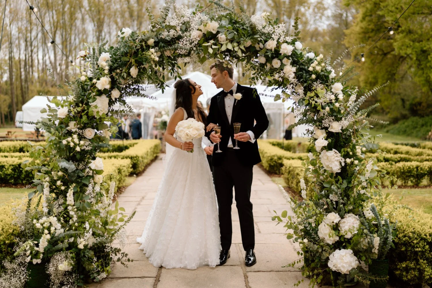Bride and groom standing under a floral arch at their outdoor wedding, holding glasses of champagne, with tents and guests in the background.