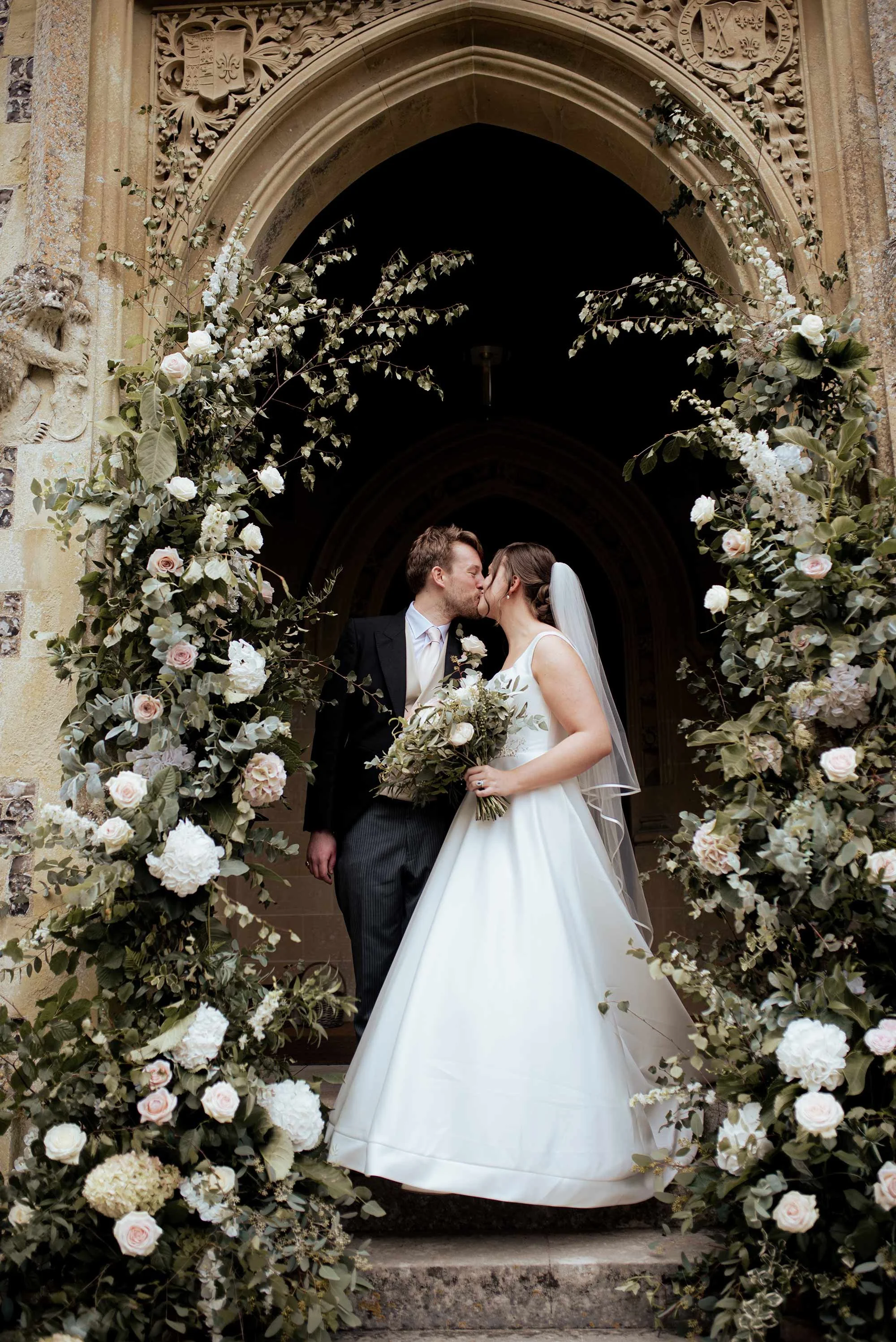 A bride and groom kiss in front of a decorated archway outside a church, surrounded by white and blush roses and greenery.