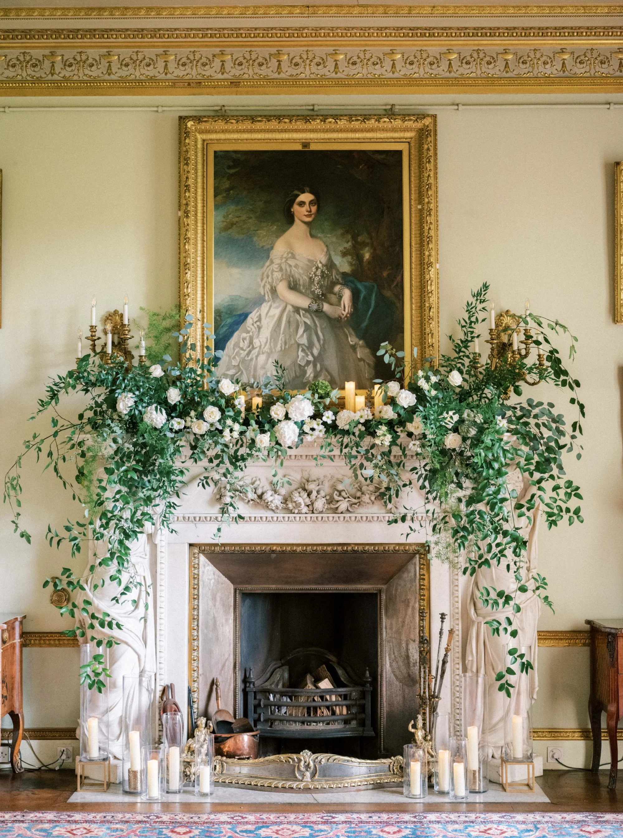 An ornate fireplace with a large classical portrait painting of a woman in a white dress hanging above it. The fireplace is decorated with lush greenery, white flowers, and numerous candles in glass containers. The room features detailed gold accents on the molding and a patterned rug on the floor.