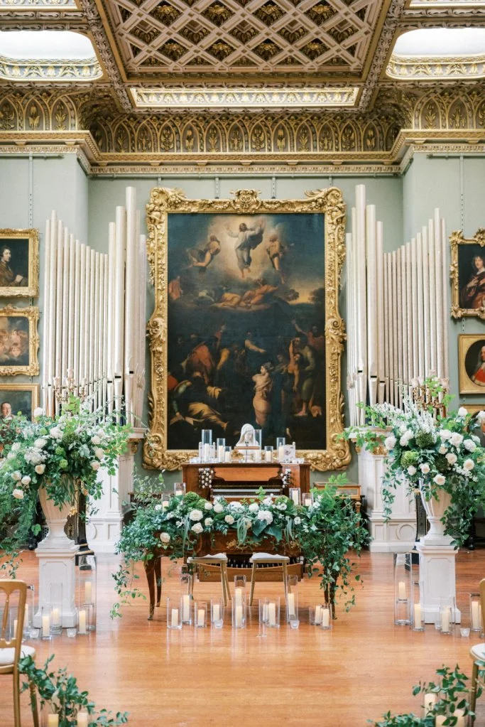 Elegant wedding altar in a historic art gallery with floral arrangements, candles, and a large Baroque painting as backdrop.