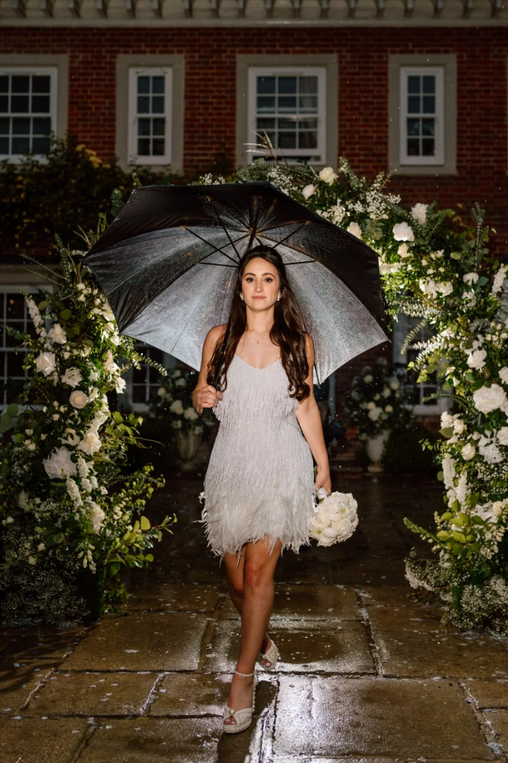 A young woman in a white dress holding a bouquet of white flowers, walking outdoors in the rain under a black umbrella, with a floral arch and brick building in the background.