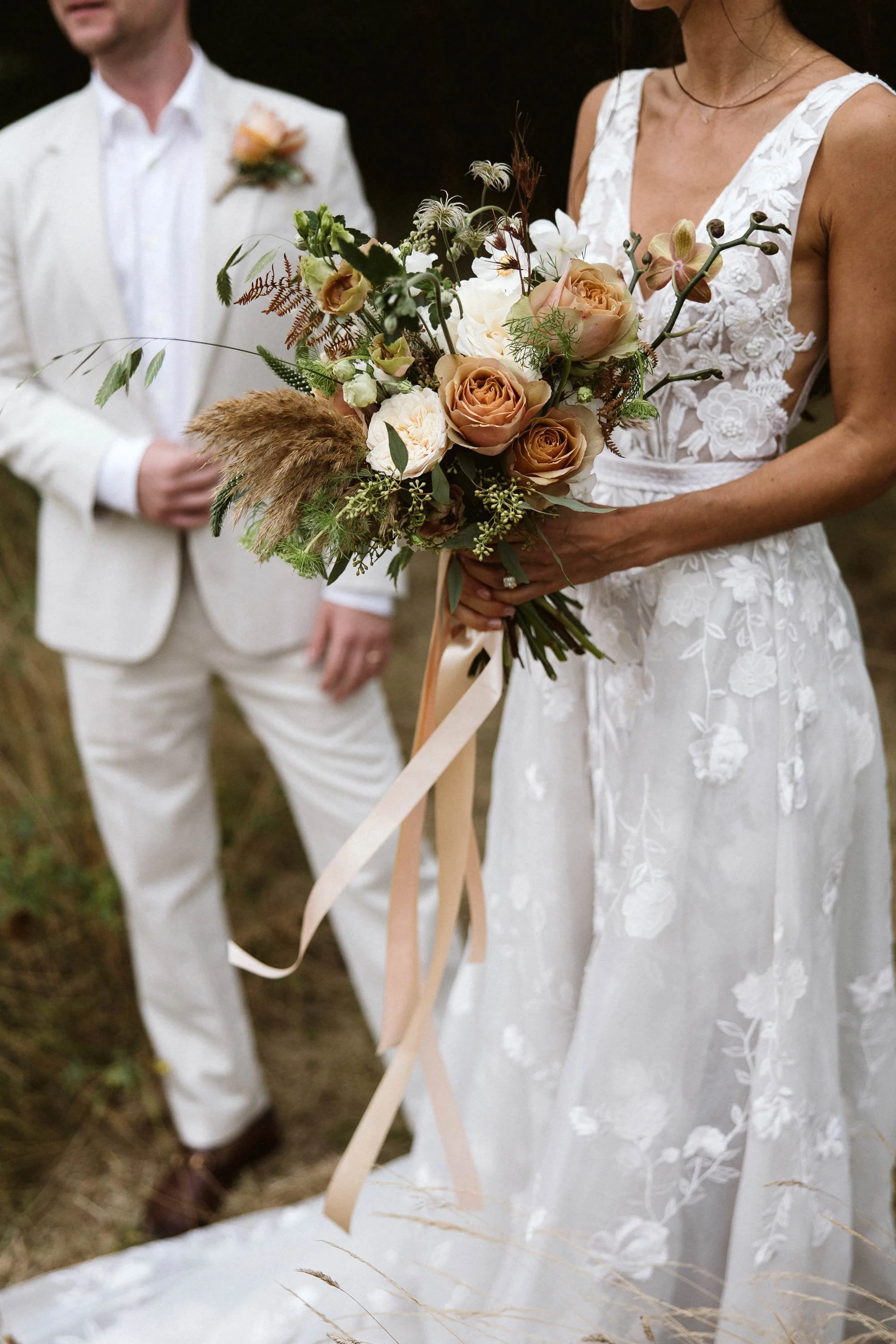 A bride wearing a white lace wedding dress holding a bouquet of flowers with roses, orchids, and greenery. A groom dressed in a cream suit with a boutonniere is standing nearby, slightly out of focus.