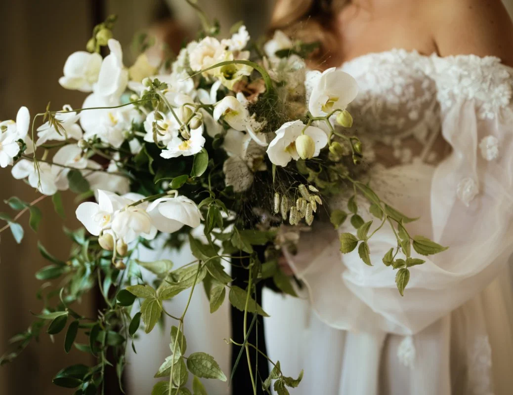 A bride holding a bouquet of white flowers and greenery, dressed in a white lace wedding gown.