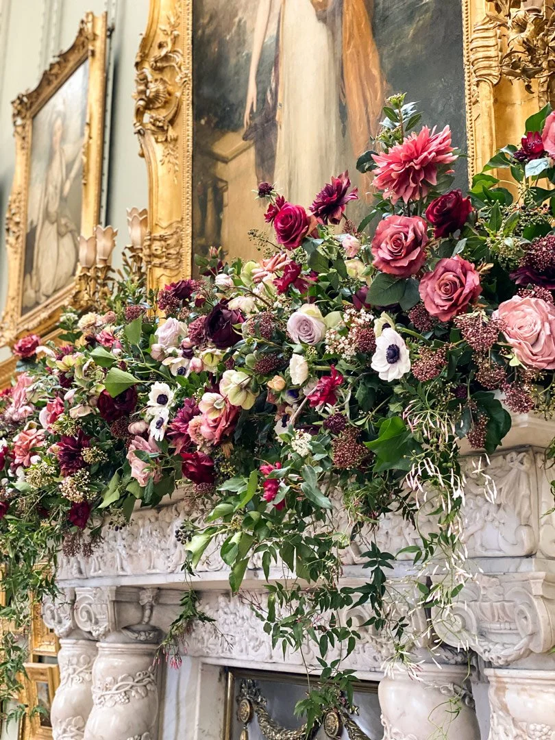 Elegant floral arrangement with pink, purple, and white flowers on a white marble fireplace in a room with ornate gold-framed paintings.