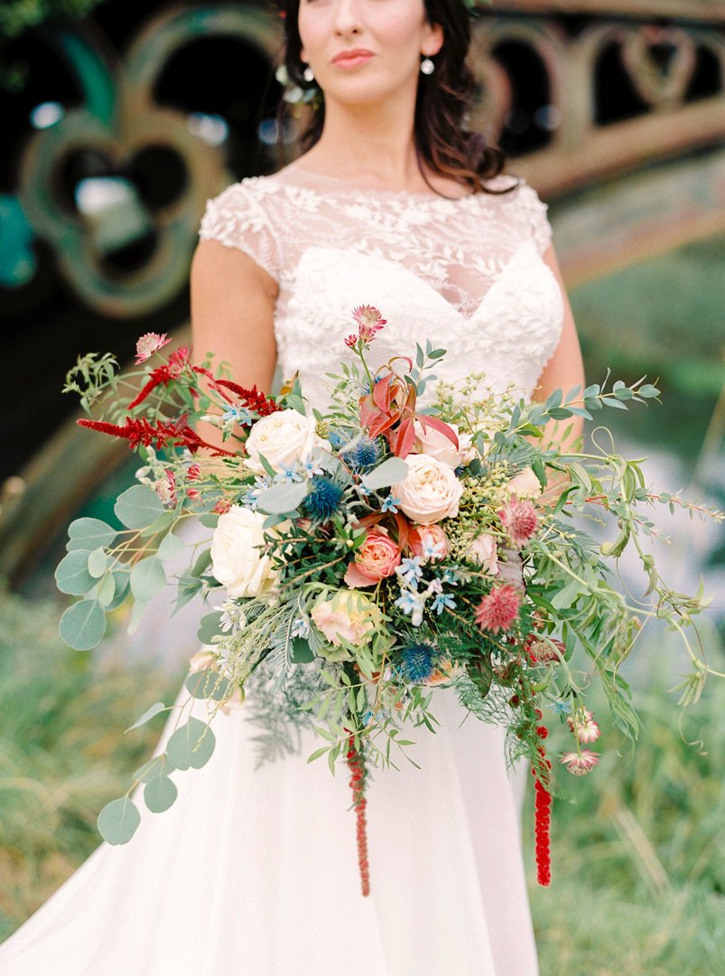 A bride holding a large bouquet of colorful flowers in front of her, with a blurred staircase in the background.