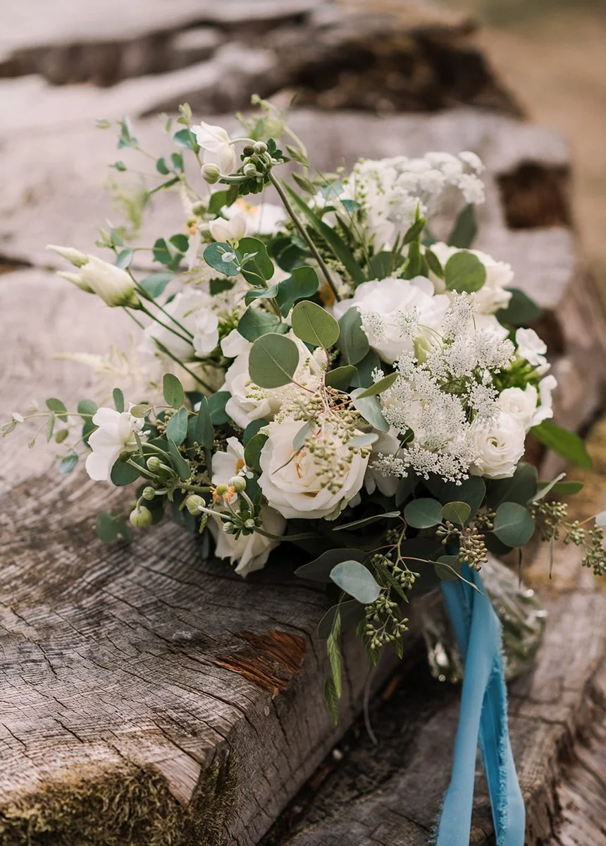 A bouquet of white flowers and green leaves resting on a rustic wooden surface, with a blue ribbon tied to it.