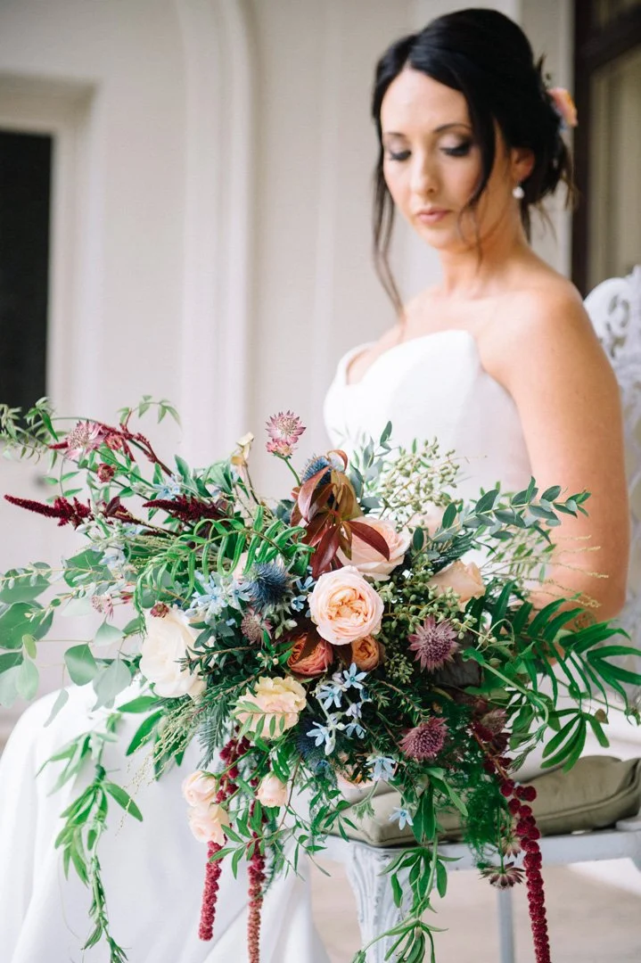 A bride in a white wedding dress holding a large, colorful bouquet of flowers with pink roses, greenery, and various wildflowers, sitting on a decorative bench in a room with light-colored walls.