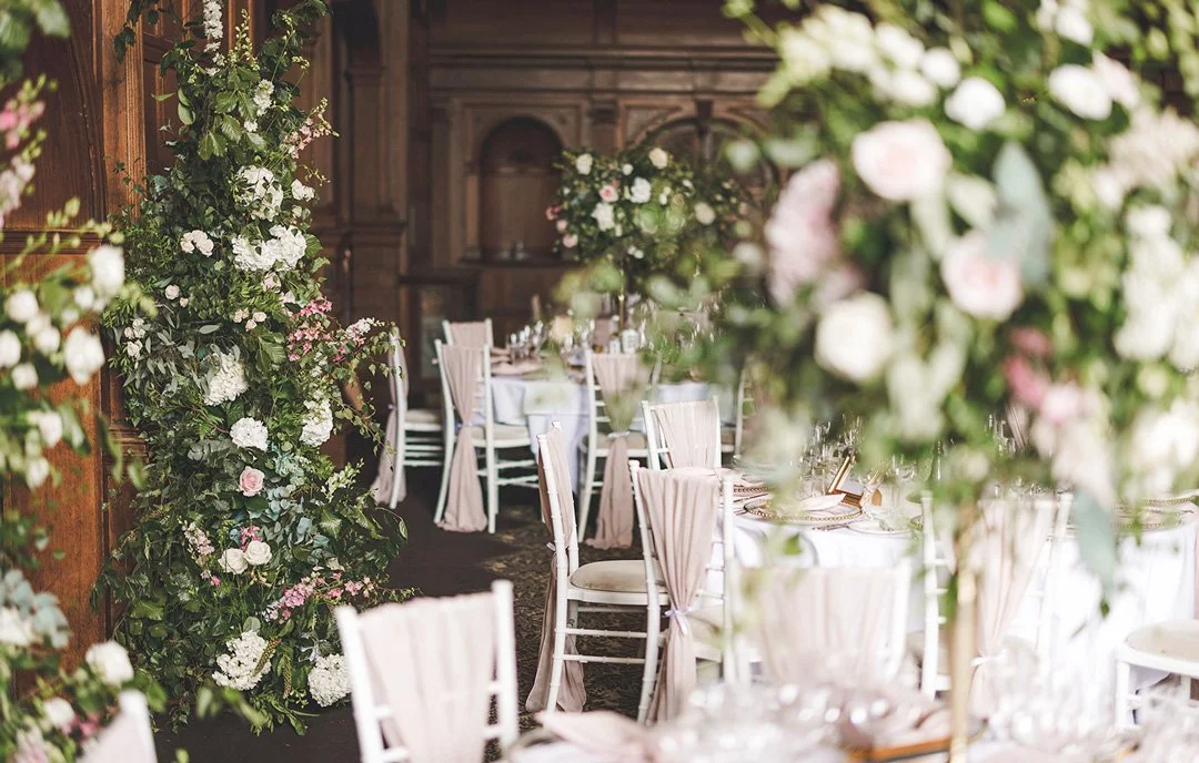 Wedding reception decorated with floral arrangements, round tables, and white chairs with pink sashes in a wood-paneled room.