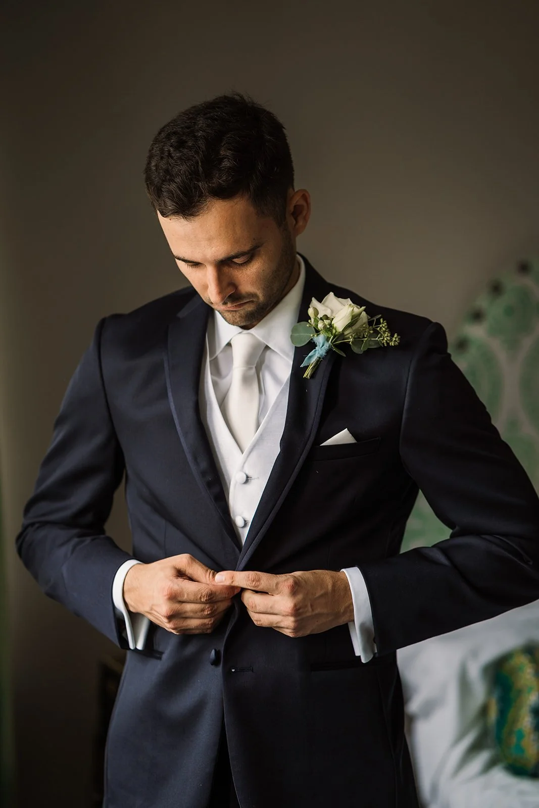 A groom in a black suit, white vest, and white tie adjusts his jacket, with a white boutonniere pinned to his lapel, standing indoors.