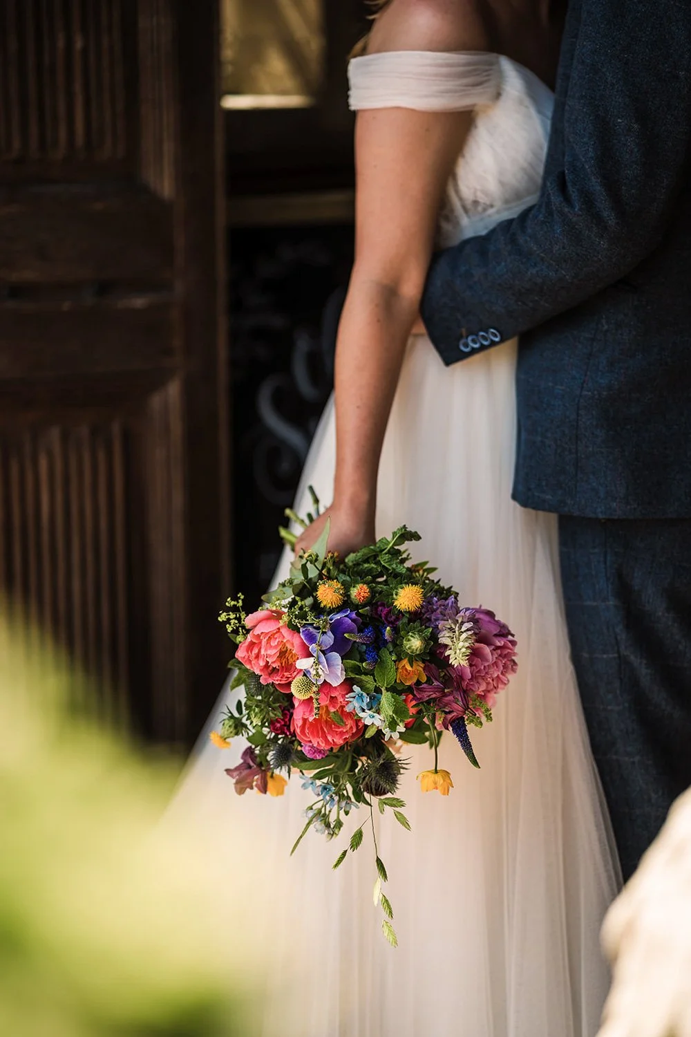 A bride holding a colorful bouquet of flowers in her right hand while standing close to a groom in a dark suit during a wedding ceremony.