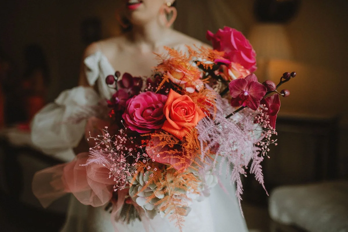 Woman holding a vibrant bouquet of pink, orange, and red flowers including roses and orchids in a warmly lit room.