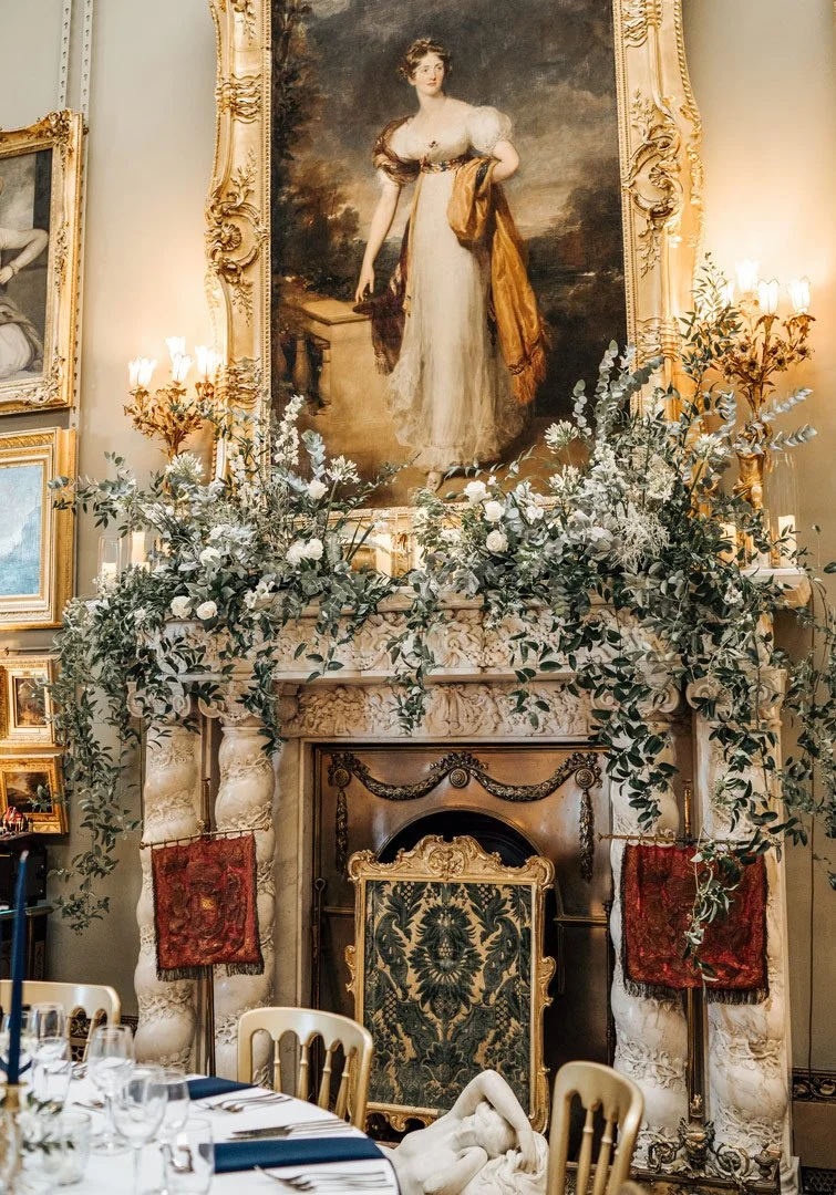 A decorated fireplace mantel with a large portrait of a woman in an elegant gown hanging above it. The mantel is adorned with beautiful wedding flowers, with ornate gold-framed mirrors and paintings on the wall around it.