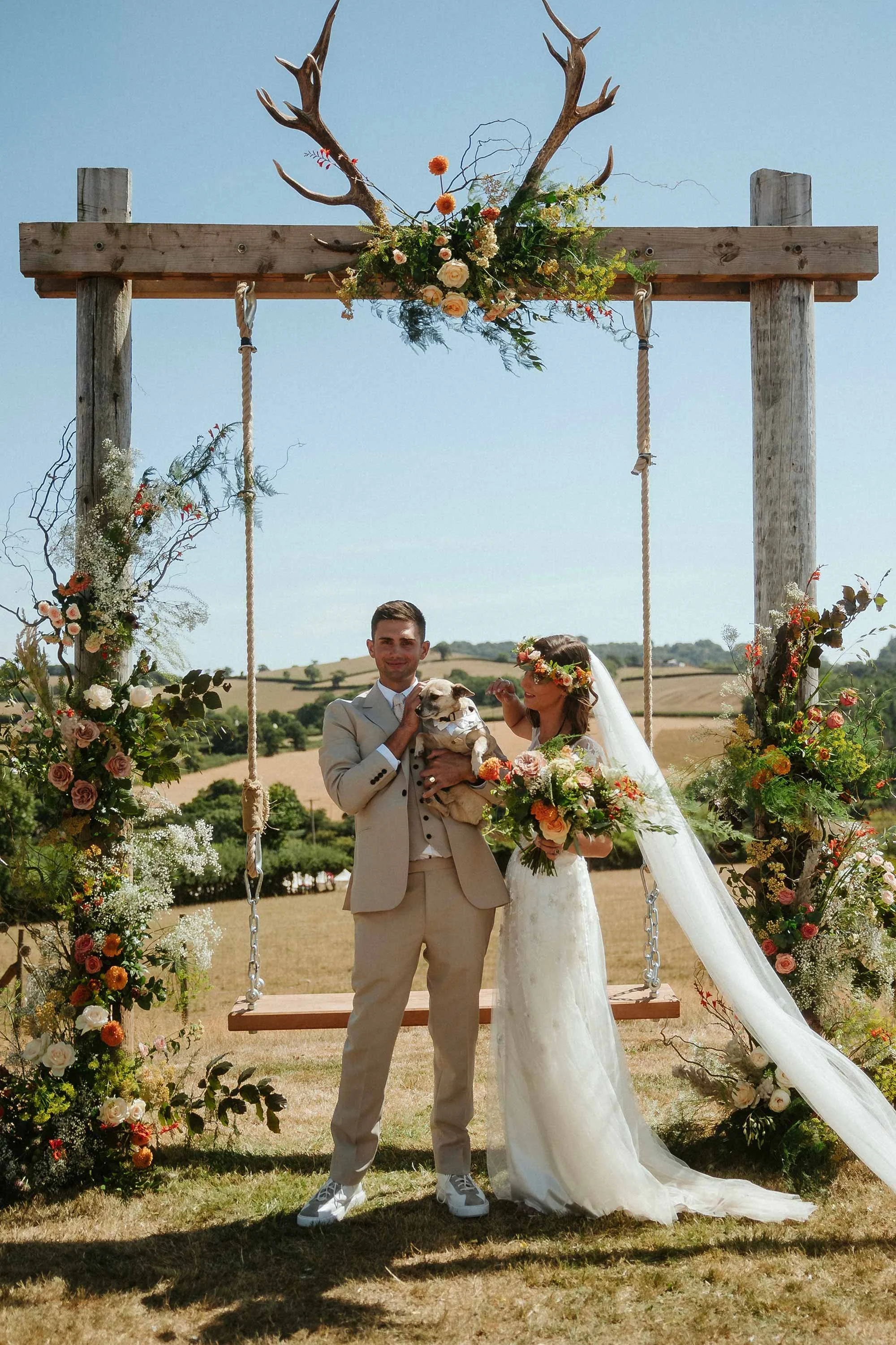An outdoor wedding ceremony with a wooden arch decorated with stunning flowers and antlers. The bride and groom are standing underneath, holding a small dog.