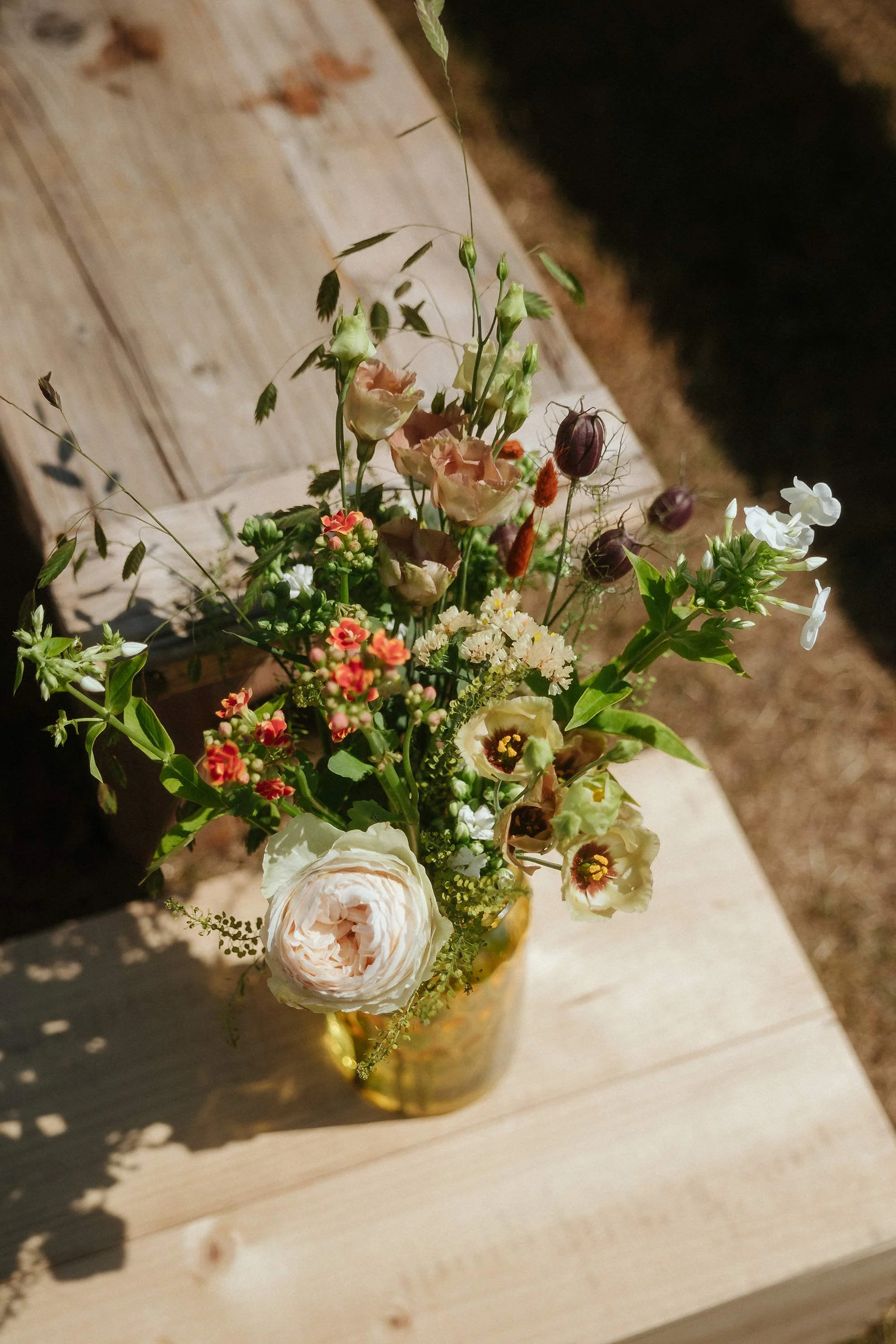 A bouquet of assorted flowers in a gold vase on a wooden surface.