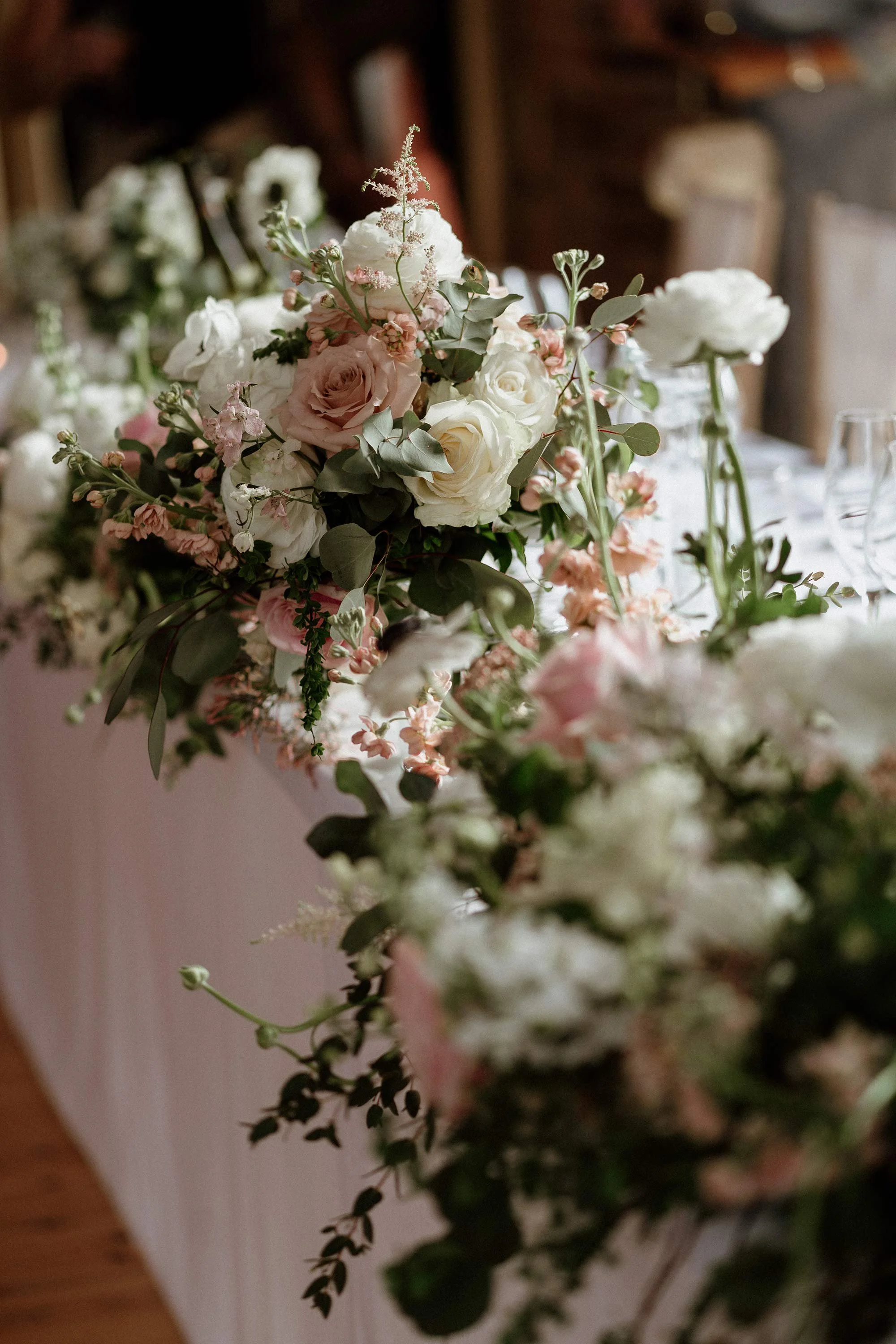 A floral centerpiece with white and light pink flowers, green foliage, and some small buds on a table.