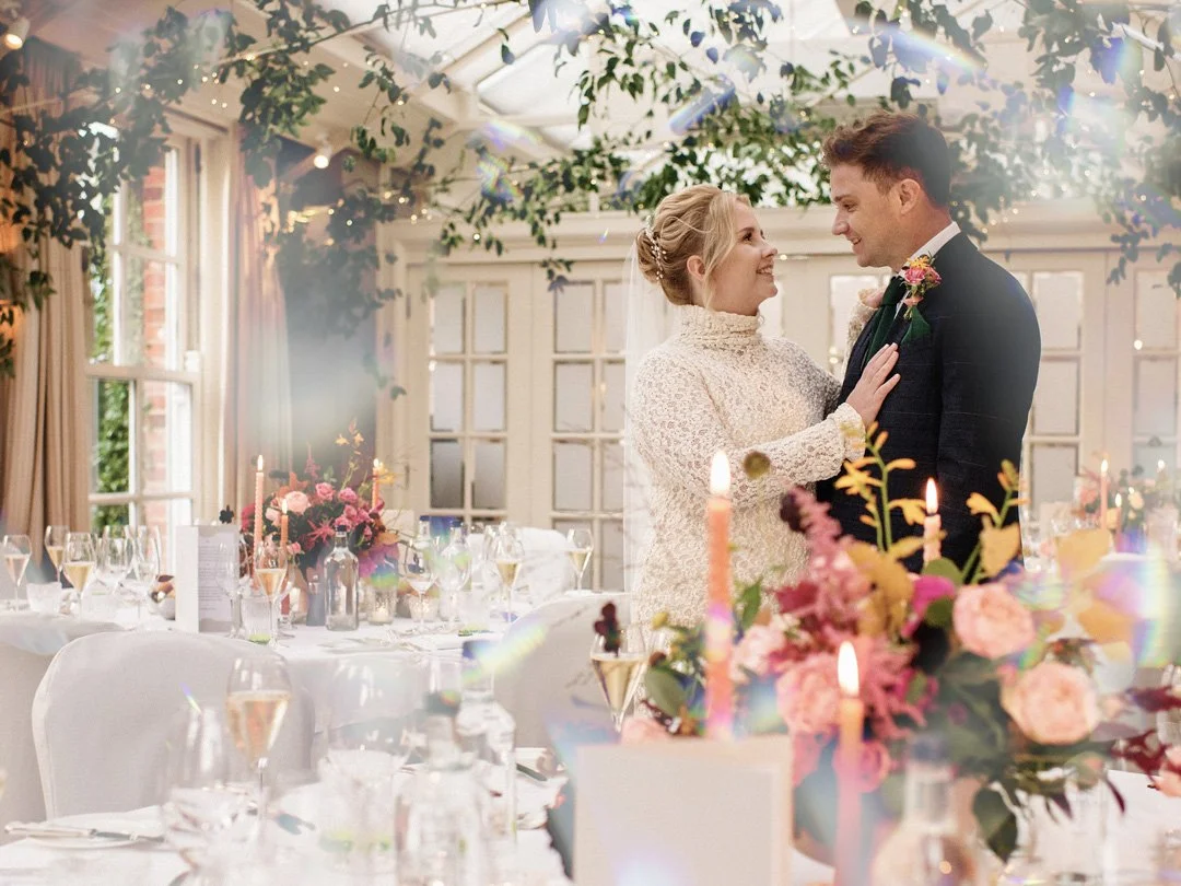 A bride and groom sharing a moment at their wedding reception, decorated with candles and floral arrangements, inside a bright, elegant venue with large windows and greenery.