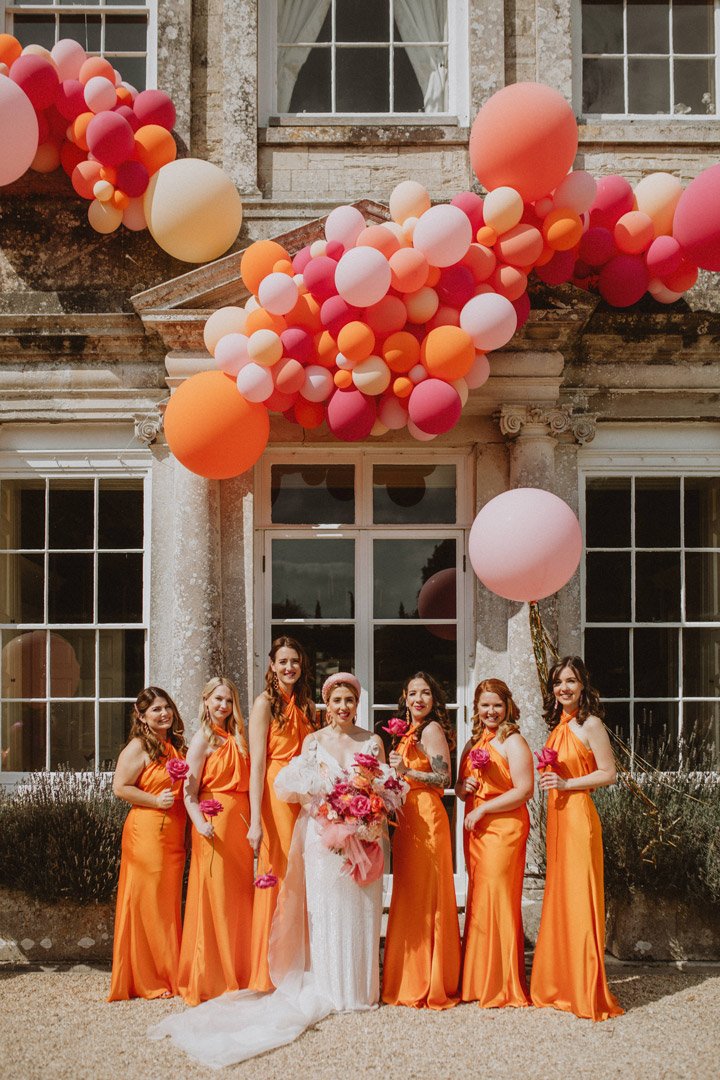Bride in white wedding dress holding a bouquet of pink flowers standing with six women dressed in matching orange bridesmaid dresses outside a building decorated with pink, peach, and orange balloons.