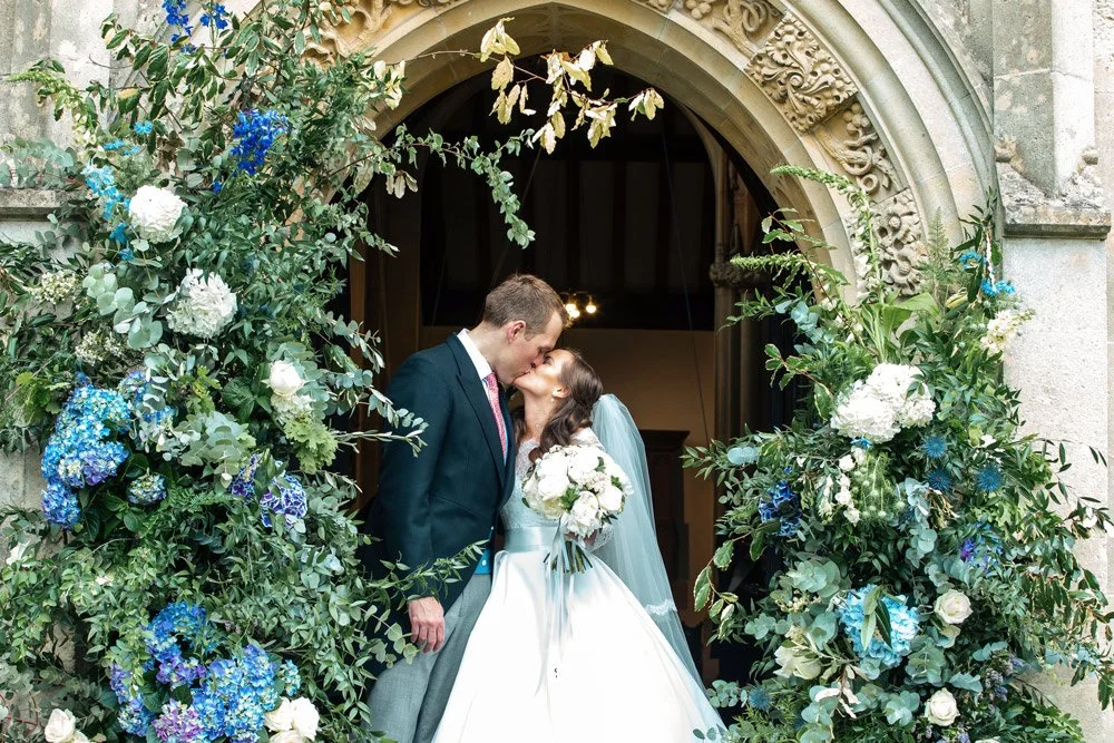 Bride and groom sharing a kiss at the altar decorated with lush greenery and white and blue flowers.