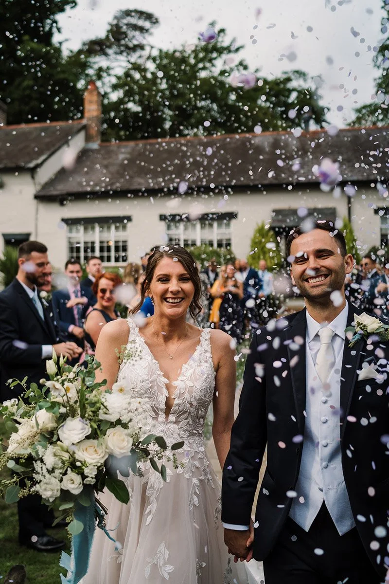 Bride and groom carrying beautiful wedding flowers smiling as confetti falls around them during their wedding celebration outdoors.