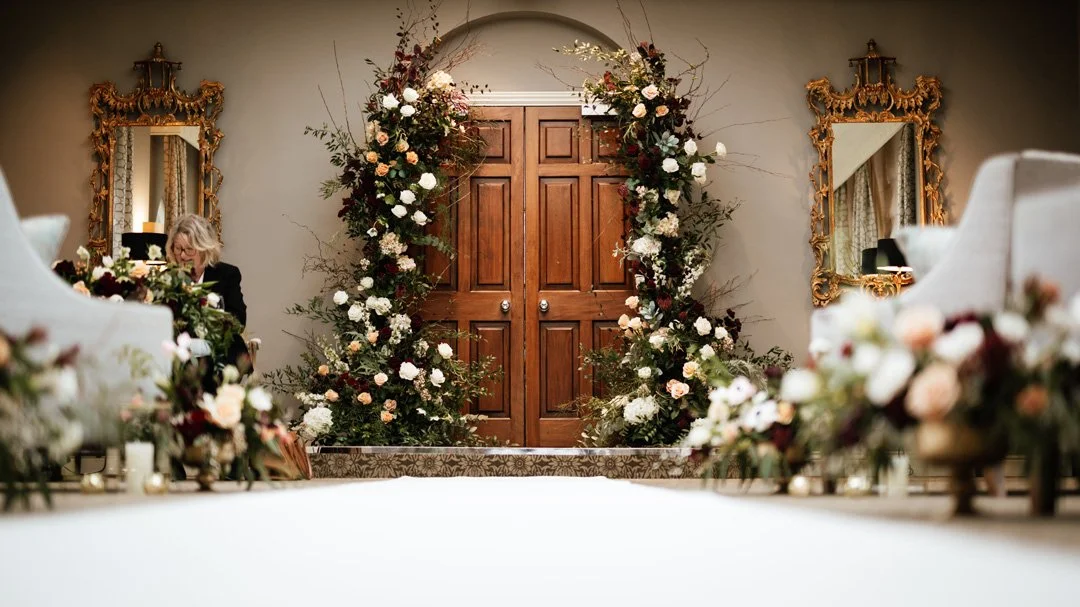 Elegant indoor setting with a large floral wedding arch in front of closed wooden double doors, flanked by gold ornate mirrors and chairs with floral arrangements.