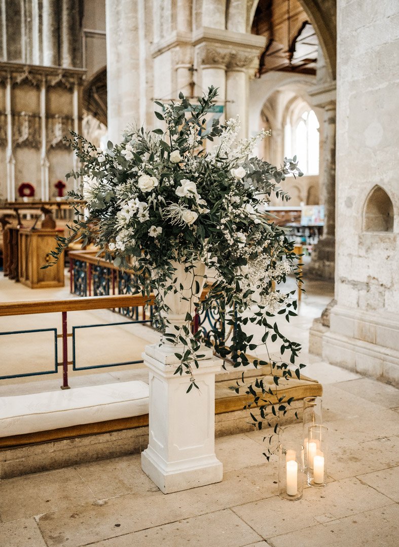 Large floral arrangement of white flowers and greenery on a white pedestal inside a church, with candles on the floor nearby.