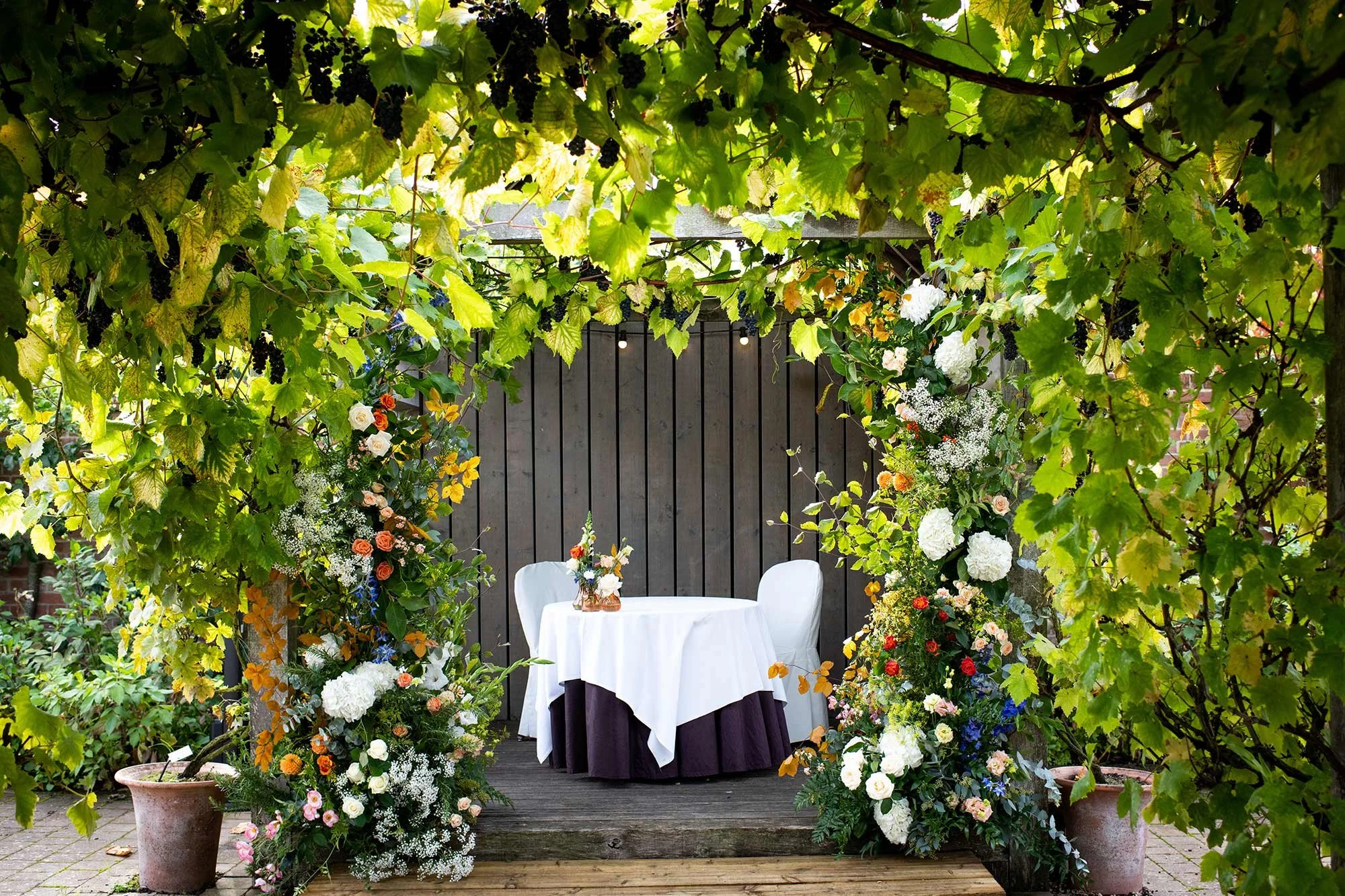 A table with a white tablecloth and a purple skirt is located outdoors, surrounded by lush green foliage and colourful flowers. There are two white chairs, and a flower arrangement in a glass vase on the table.